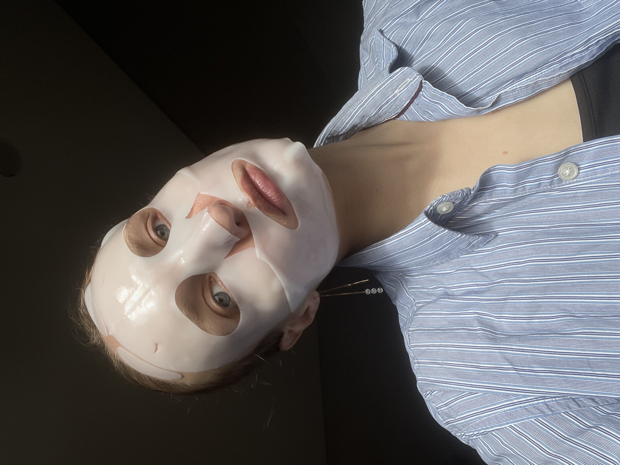 A close-up, eye-level portrait features a person wearing a white, translucent sheet face mask. The mask covers the entire face, with cutouts revealing the person's light blue or gray eyes, nose, and slightly parted lips. The eyes are open and gaze directly at the viewer with a neutral expression. Light-colored hair is visible around the edges of the mask.
The person is wearing a light blue and white striped button-down shirt, which is partially unbuttoned, revealing a dark garment beneath and a small area of the chest. A decorative earring or ear cuff, featuring three small, reflective beads, is visible on the right ear.
The background is uniformly dark and out of focus, suggesting an indoor setting with subdued lighting. No specific environmental details or text are visible. The image captures a moment of personal care, likely a skincare routine, and is set in Подшивалово, Russia.