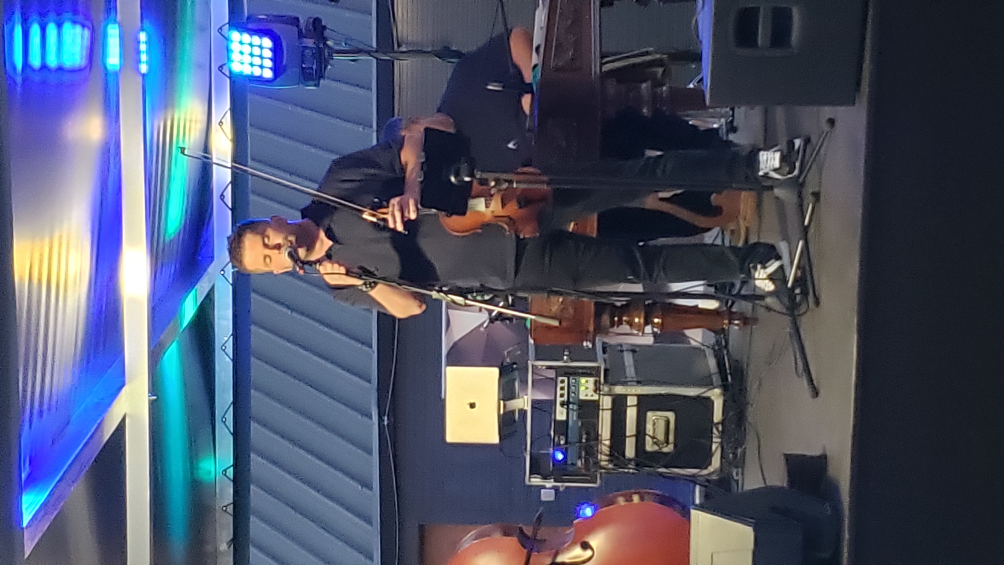 A male performer, dressed in a black polo shirt and black pants, stands on a stage, holding a violin and bow, while singing into a microphone. He has closed eyes and his head is tilted slightly upwards. He wears black and white sneakers. To his left, another person, possibly a performer, is seated behind a dulcimer. In the background, there is audio equipment, including speakers and racks of electronics, and a laptop on a stand. Stage lighting illuminates the scene with blue and white lights. The backdrop appears to be a dark corrugated metal wall. This event is taking place in Prešov, Slovakia, possibly at an outdoor venue or a venue with an open-air roof structure, suggested by the lighting and the visible metal roofing.