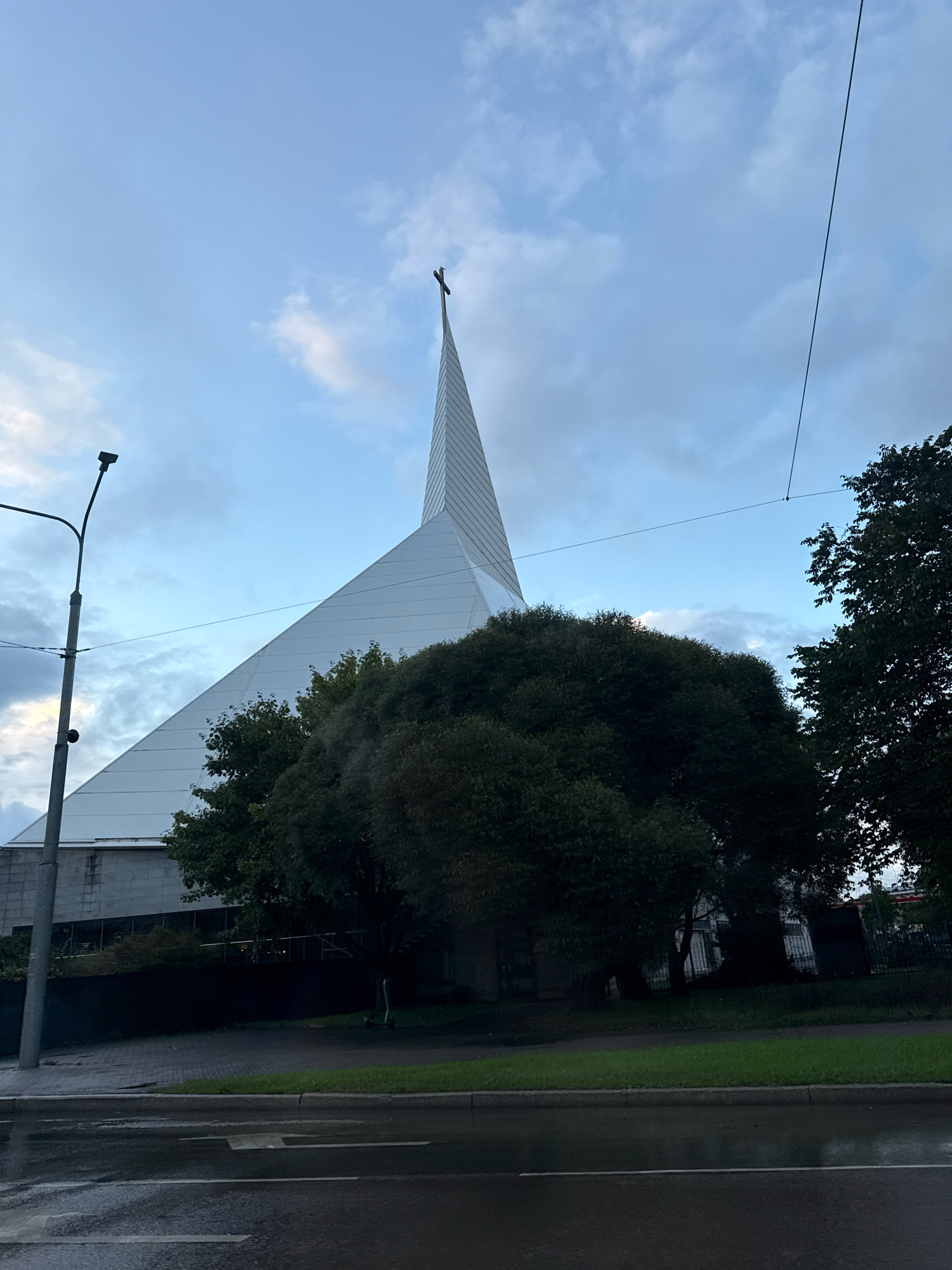 The image captures a modern church building with a distinctive, angular white spire topped with a cross, set against a cloudy sky. The church, likely the Saint Peter and Saint Paul Cathedral in Tallinn, Estonia, features a white, sloped roof that tapers to a sharp point. In the foreground, a wet asphalt road with white lane markings suggests recent rain. A grey concrete sidewalk runs alongside the road, bordered by a strip of green grass. Behind the sidewalk, a large, dark green tree with dense foliage obscures the lower portion of the church. A lamppost stands to the left of the church. Power lines are visible stretching across the sky.