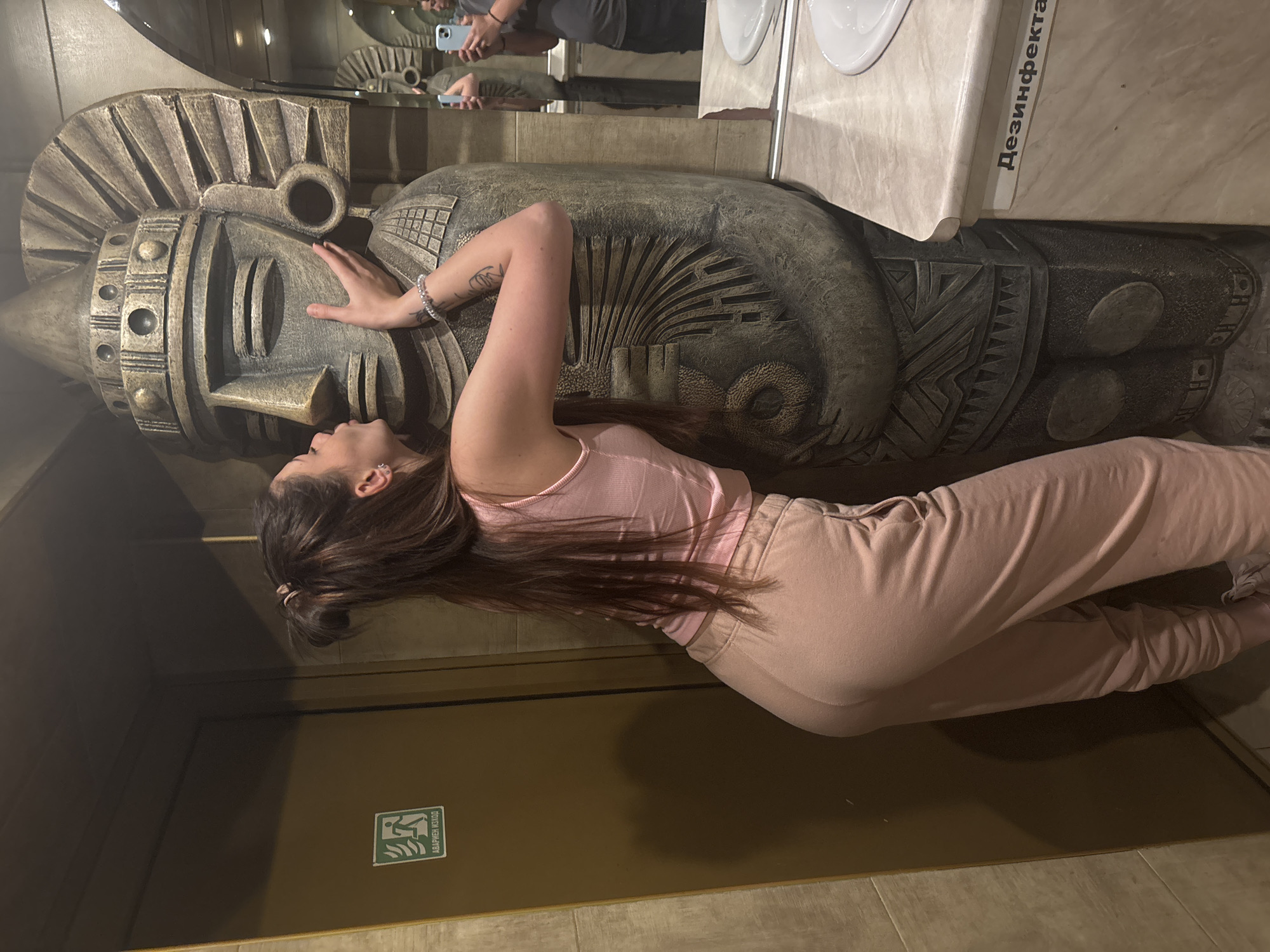 A woman is leaning against a large, stylized stone sculpture of a figure wearing a headdress inside a building in Sofia, Bulgaria. The woman is wearing a pink tank top and sweatpants. Her left hand is touching the sculpture's face. The sculpture is positioned in front of a countertop with a sink. The word "Дезинфекта" is visible in Cyrillic on the countertop. In the background, a mirror reflects the scene, including a person holding a phone. An emergency exit sign is present on a door to the left of the sculpture. Tiled flooring is observed in the image.