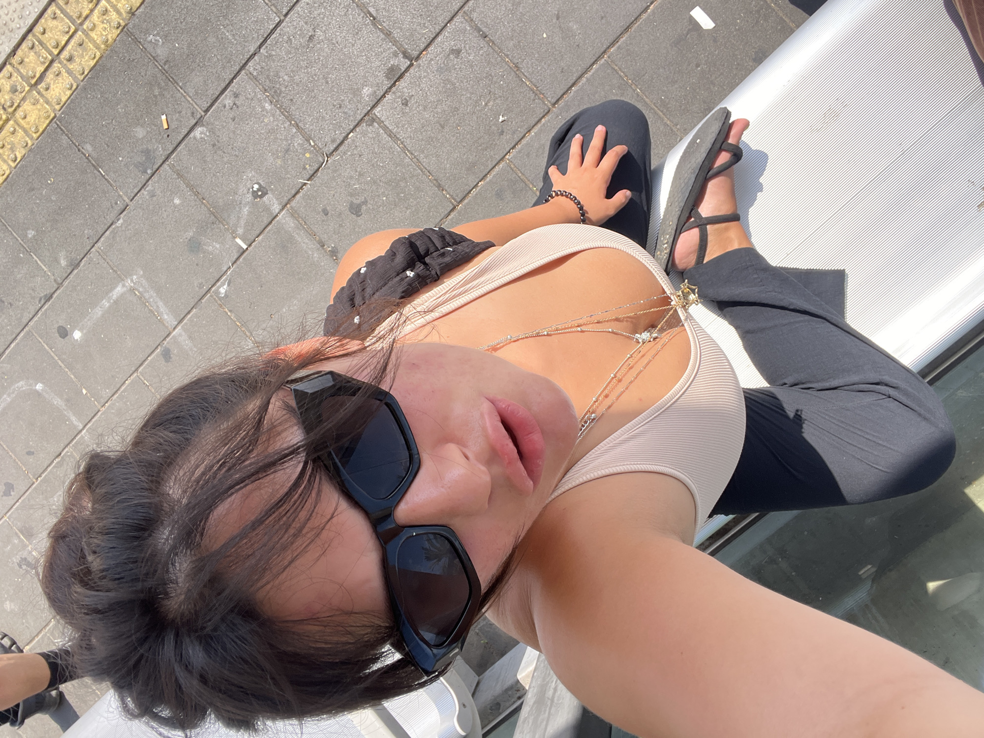 A high-angle, medium-shot selfie features a young woman with dark hair pulled back, wearing large, black, angular sunglasses, a light-colored ribbed tank top, and dark leggings. She is positioned with her head tilted to the side and her mouth slightly open. Multiple necklaces with pendants are visible on her chest. Her right hand rests on her leg, which is adorned with a dark beaded bracelet. Her left foot, wearing a black flip-flop, is visible at the bottom right of the frame. The background consists of grey paving stones with textured yellow tactile paving bricks along the top edge, suggestive of a public pedestrian area, possibly near a street or sidewalk in Kiryat Motzkin, Israel. A discarded cigarette butt is visible on the paving stones. The woman is seated on a white, ridged surface, possibly a bench or a piece of street furniture, casting a shadow.