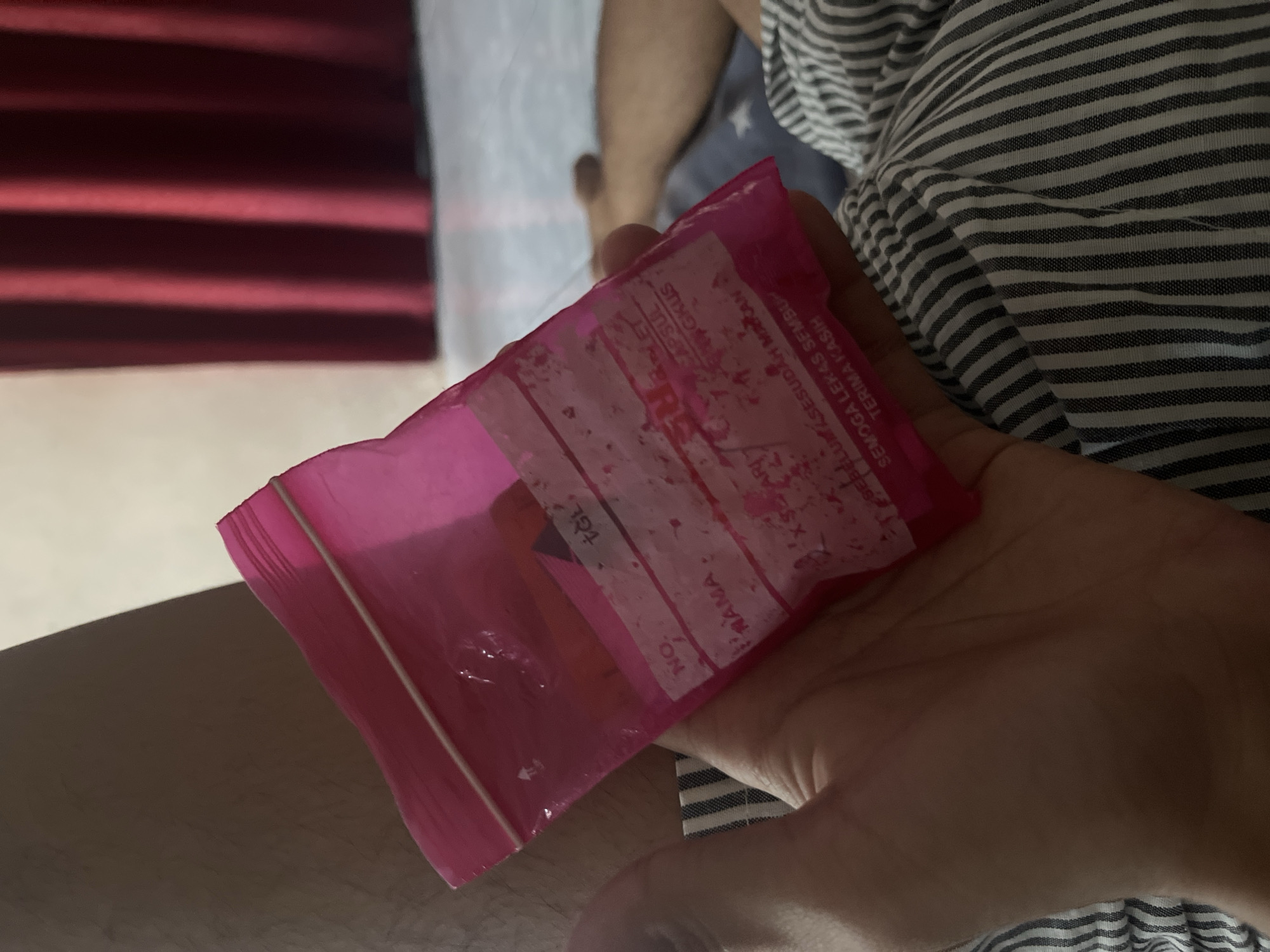 The image shows a person's hand holding a pink plastic bag containing what appears to be medication. The bag has labels with text in Indonesian, including fields for "NO.", "NAMA" (name), and a date ("TGL"). Text below indicates instructions related to medication. The background is blurred, showing part of a person's leg and striped fabric, possibly bedding. Red curtains are visible in the background, suggesting an indoor setting. The image is taken from a close-up perspective, focusing on the hand and the bag. The scene is likely in a residential setting within Paweden, Indonesia.