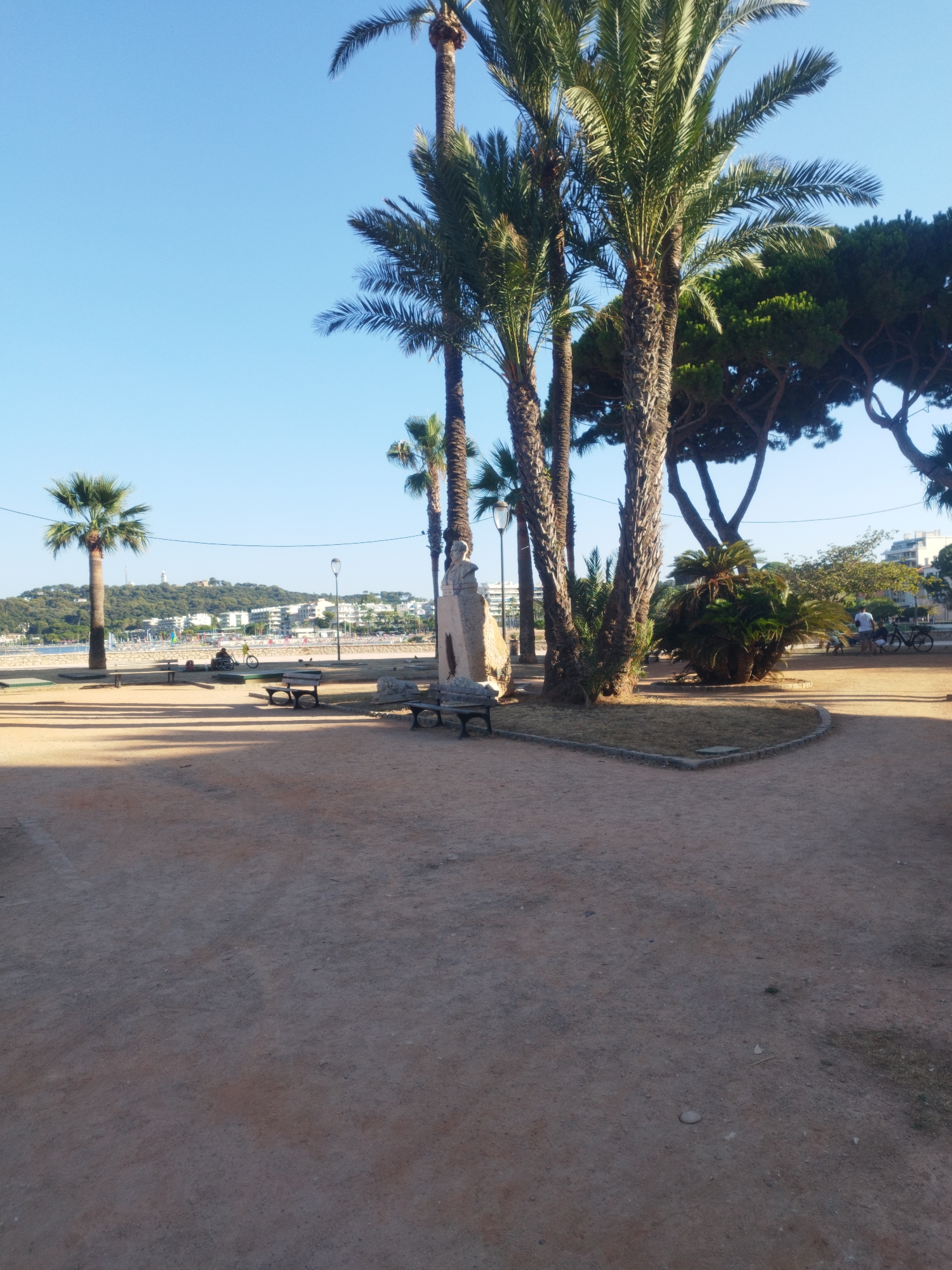 The image depicts a sunlit park area in Antibes, France, with the Mediterranean Sea and cityscape in the background. Several palm trees and larger trees are prominent in the foreground and midground, casting shadows on the sandy ground. In the midground, there are two park benches. To the right of the benches, a stone monument with a bust is visible. Further back, beyond a low barrier, a beach is apparent with umbrellas and a few scattered people. The cityscape of Antibes rises behind the beach, with buildings and a hill topped by a white structure. Two individuals are seen in the distance on the beach area; one is riding a bicycle and another is seated. In the far right of the midground, a group of people are standing near a bicycle, and one person is walking away from the camera. The sky is clear and blue.