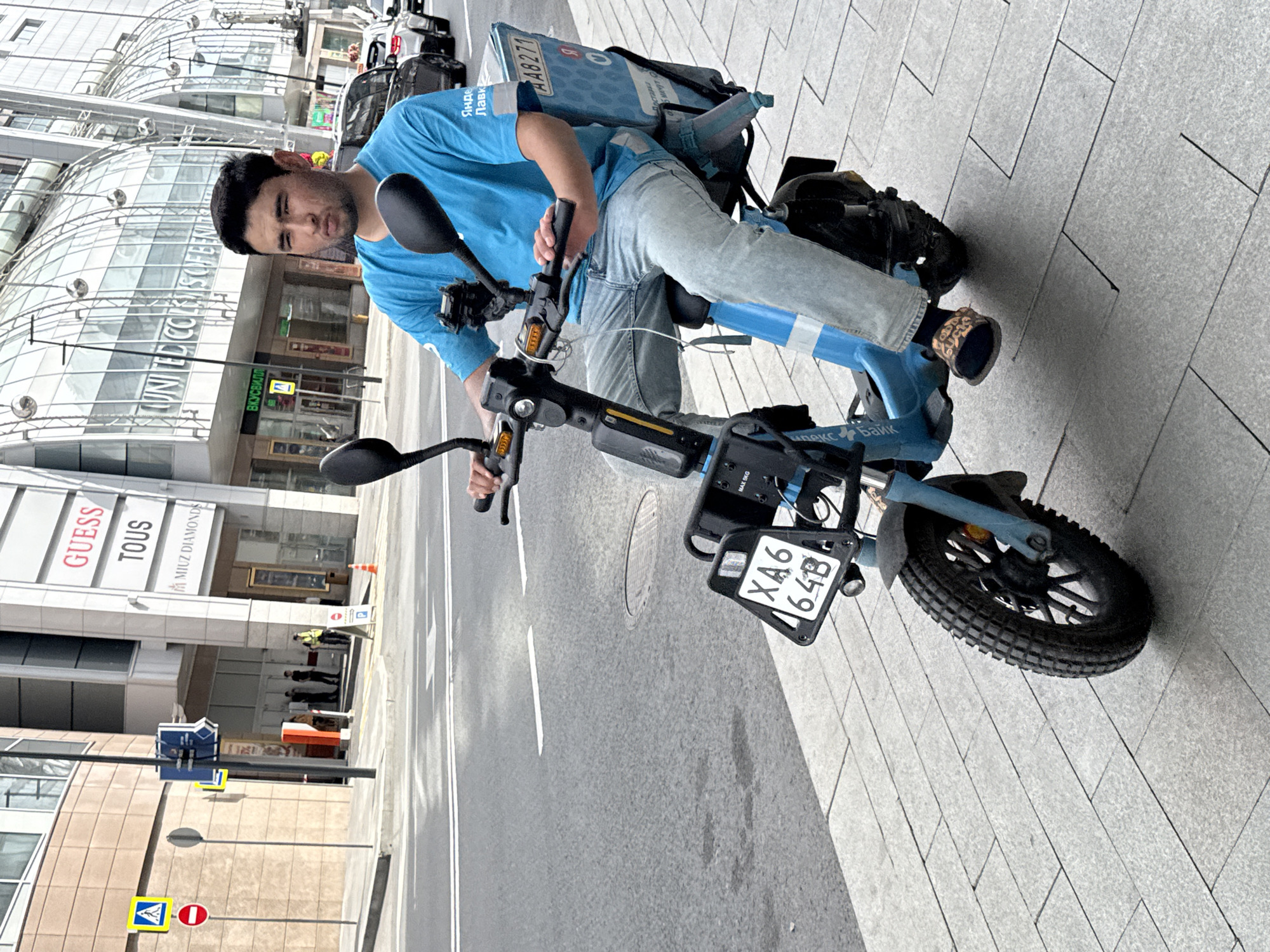 A young man wearing a blue t-shirt and light blue jeans is riding a blue electric scooter on a sidewalk in Moscow, Russia. He has dark hair and is looking straight ahead with a neutral expression. The scooter has a large blue delivery box on the back with Russian text and a license plate that reads "XA6 64B". The background shows a modern building with glass windows and signage, including "GUESS" and "TOUS". There are also traffic signs and streetlights visible. The sidewalk is paved with light-colored rectangular tiles.