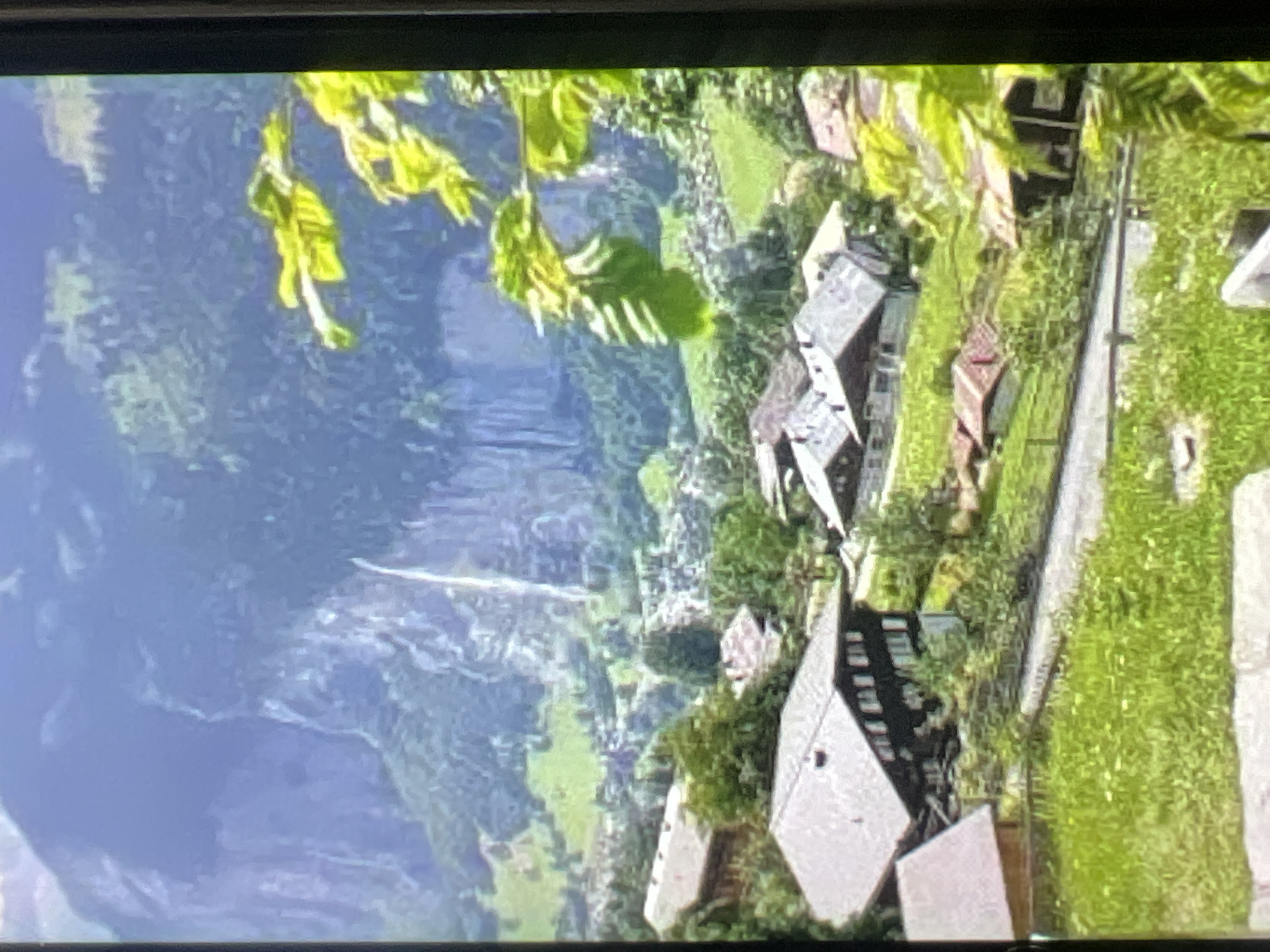 This is an aerial or high-angle shot of a picturesque mountain village, likely in the Alps. The scene is dominated by steep, verdant green mountainsides and a dramatic waterfall cascading down a rocky cliff face. Nestled within the valley is a charming village composed of traditional chalets with dark wooden roofs and stone accents. The architecture suggests a European alpine setting.
The village is situated along a winding path or road, with several buildings visible. Some structures appear to be residential homes, while others might be inns or other village amenities. The surrounding landscape is lush with trees and grass, indicating a temperate season, likely summer or late spring, given the vibrant green foliage.
In the foreground, green leaves from a tree branch hang into the frame, adding a natural, framing element to the view. The lighting suggests it is daytime, with good visibility, though no specific time of day can be determined. There is no visible text in the image. The overall impression is one of tranquility and natural beauty, characteristic of a secluded mountain resort. No people are visible in the frame, focusing the attention entirely on the landscape and the village architecture. The scene evokes a sense of peace and the charm of rural life in a stunning natural environment.