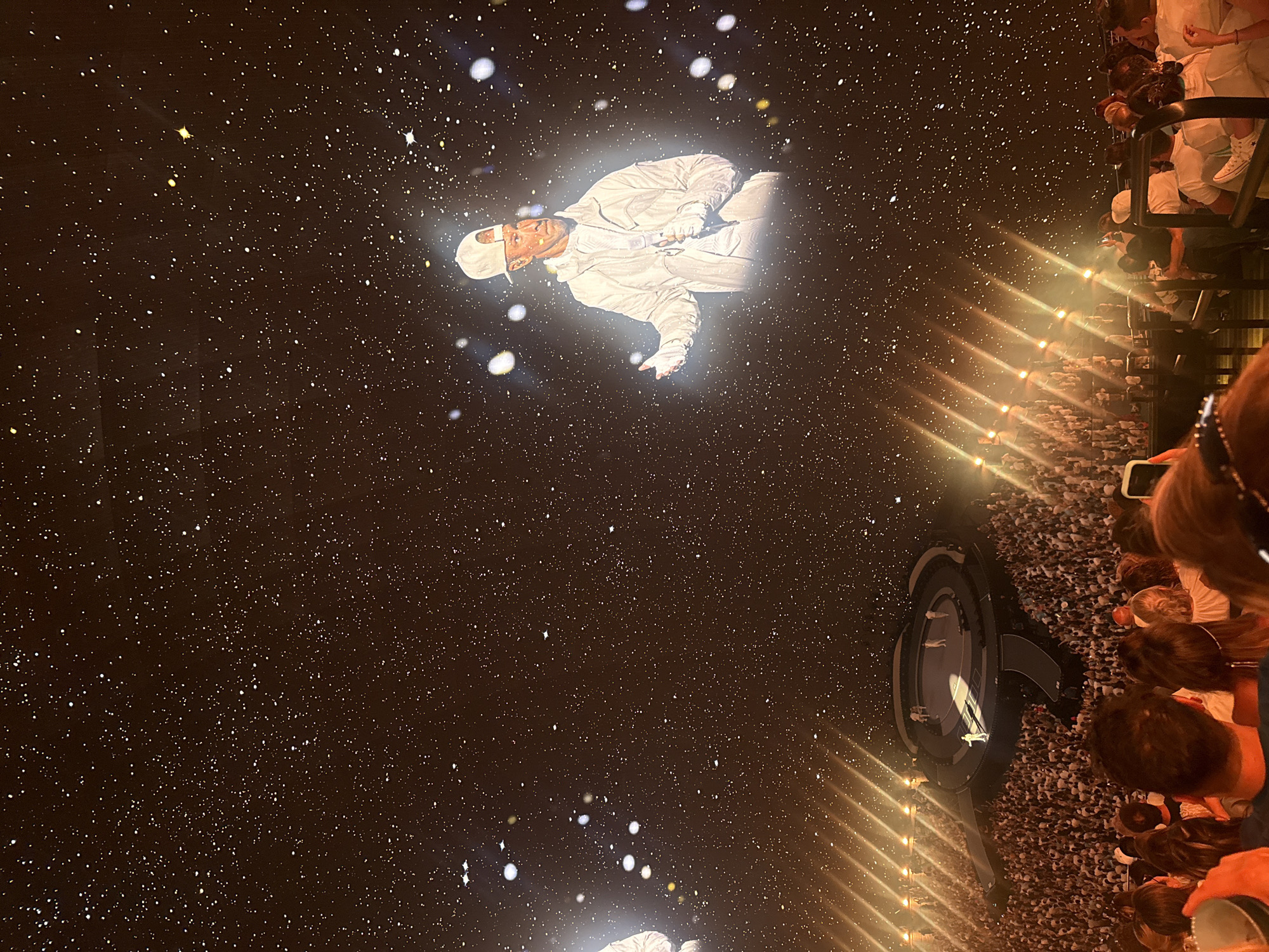 The image shows a large concert venue, likely in Paradise, United States of America, illuminated by a projection of a person in a white hoodie and baseball cap against a dark, star-filled background. The projection is centered on the right side of the frame. In the foreground and middle ground, a massive audience, mostly clad in white, is seated, facing a circular stage. On the stage, several figures are visible, one of whom appears to be performing. The overall ambiance is that of a live concert event with extensive lighting and a starry projection creating a celestial atmosphere.