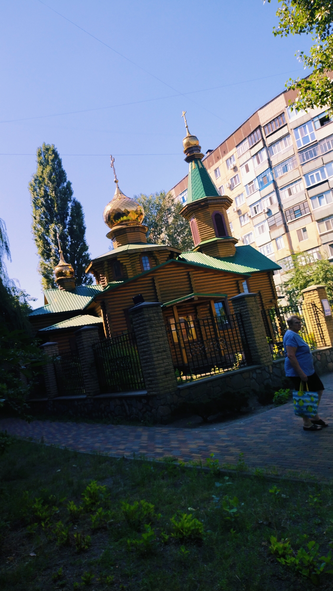 In an urban setting, a low-angle, slightly tilted shot captures a wooden Orthodox church with golden domes on the left, juxtaposed against a multi-story apartment building on the right. The church features green roofing and a fence surrounding its perimeter, with a person walking on a paved path in front of it. The sky is clear and blue. The scene is lit by sunlight, casting shadows on the ground.