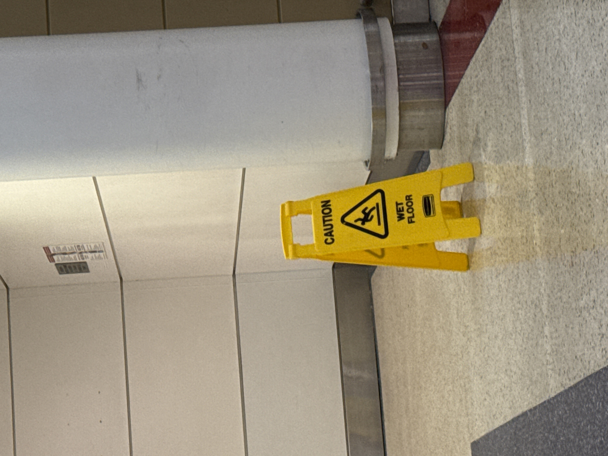 The image depicts a yellow "Caution Wet Floor" sign situated on a tiled floor next to a wall and a large white cylindrical object. The sign warns of a slippery surface with a graphic of a person falling. The environment appears to be indoors, potentially a public space like a commercial building or a transportation hub. Based on the interior design and the presence of the sign, it is likely that this photo was taken at Chicago O'Hare International Airport.