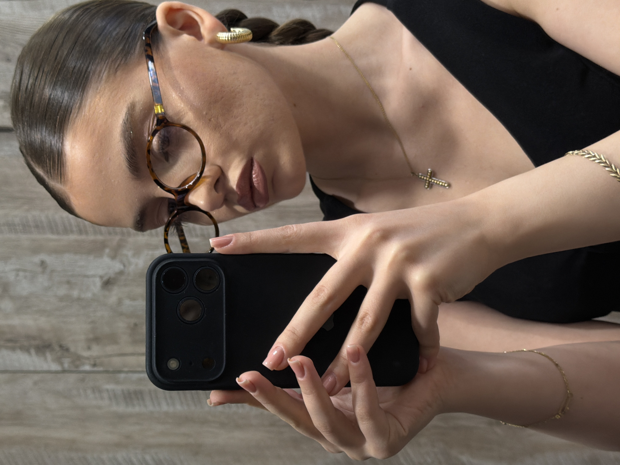 A young woman is depicted in a close-up, rotated portrait, appearing to take a selfie or record herself with a smartphone. Her dark hair is neatly pulled back, revealing a braid near her right ear. She wears large, round tortoiseshell glasses and a gold-toned ridged hoop earring. A delicate gold chain with a small, ornate cross pendant adorns her neck, complementing a gold-toned woven bracelet on her right wrist and a thinner chain bracelet on her left arm. Her lips are painted with a subtle nude/pink shade.
She holds a black smartphone horizontally in both hands, its back showing a prominent three-lens camera system and the faint outline of an Apple logo. Her fingernails are manicured with a natural-toned polish. The background is a light-colored wooden surface with visible grain, suggesting an indoor setting. The image is presented rotated 90 degrees clockwise. The location context provided for this image is Yerevan, Armenia.