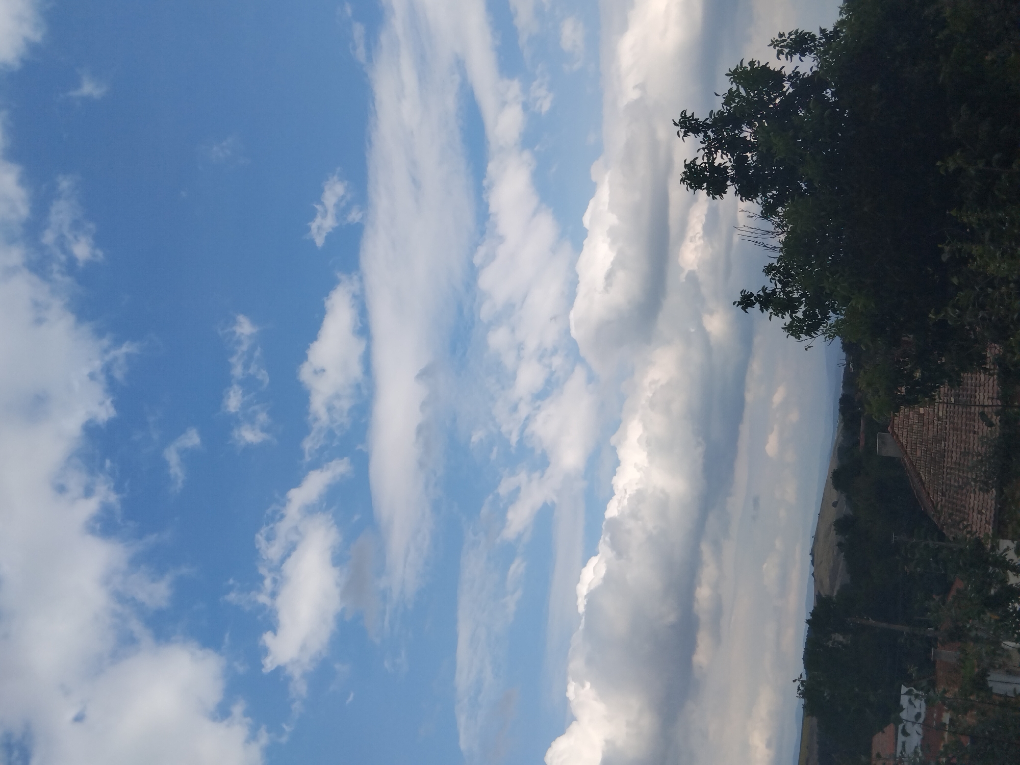 The image depicts a rural landscape under a partly cloudy sky. In the foreground, the tops of trees and parts of rooftops are visible, suggesting a residential area. The sky is predominantly blue with scattered white and grey clouds. In the midground, a dense line of trees separates the residential area from rolling hills in the distance. The hills are covered in vegetation and appear to be agricultural or natural landscapes. The overall impression is a serene, natural environment with signs of human habitation. The image does not contain any people, actions, or interactions.