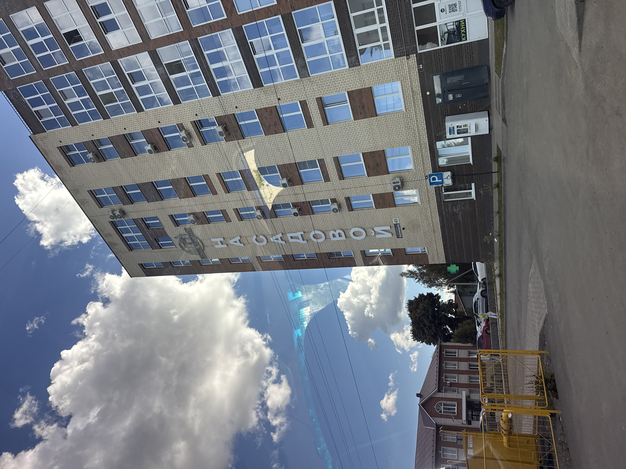 The image depicts a building with the Cyrillic inscription "НАСАДОВОИ" (NASADOVOI) in Mozhga, Russia. The building is of light brick with brown brick accents around the windows. Air conditioning units are visible beneath some of the windows. A parking sign with a wheelchair symbol is present. A kitchen store is located in the building.
A blue sky with scattered white clouds is visible above and beside the building. Wires stretch across the sky. In the foreground, a yellow metal barrier and a section of road are visible. Across the road, is another building.
Cars are parked along the side of the street.