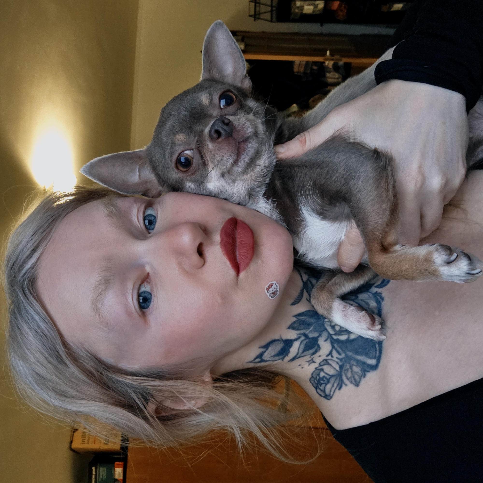 A portrait of a person with light blonde hair and blue eyes cuddling a small gray and white Chihuahua. The person has bright red lipstick and a Hello Kitty sticker on their cheek, along with a large floral tattoo on their upper chest. The background is blurry, but appears to be an interior space with warm lighting. The location is not discernible from the image.