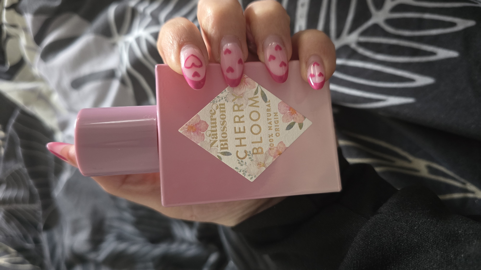 A hand with manicured nails featuring pink tips and heart designs is holding a rectangular pink bottle labeled "Nature Blossom Cherry Bloom 100% Natural Origin." The background is a patterned grey and white fabric, with a small portion of a black garment visible in the lower right. The image was likely taken indoors, but the specific location within Zirndorf, Germany, cannot be determined from the image. No specific actions or interactions are discernible beyond the hand holding the bottle.