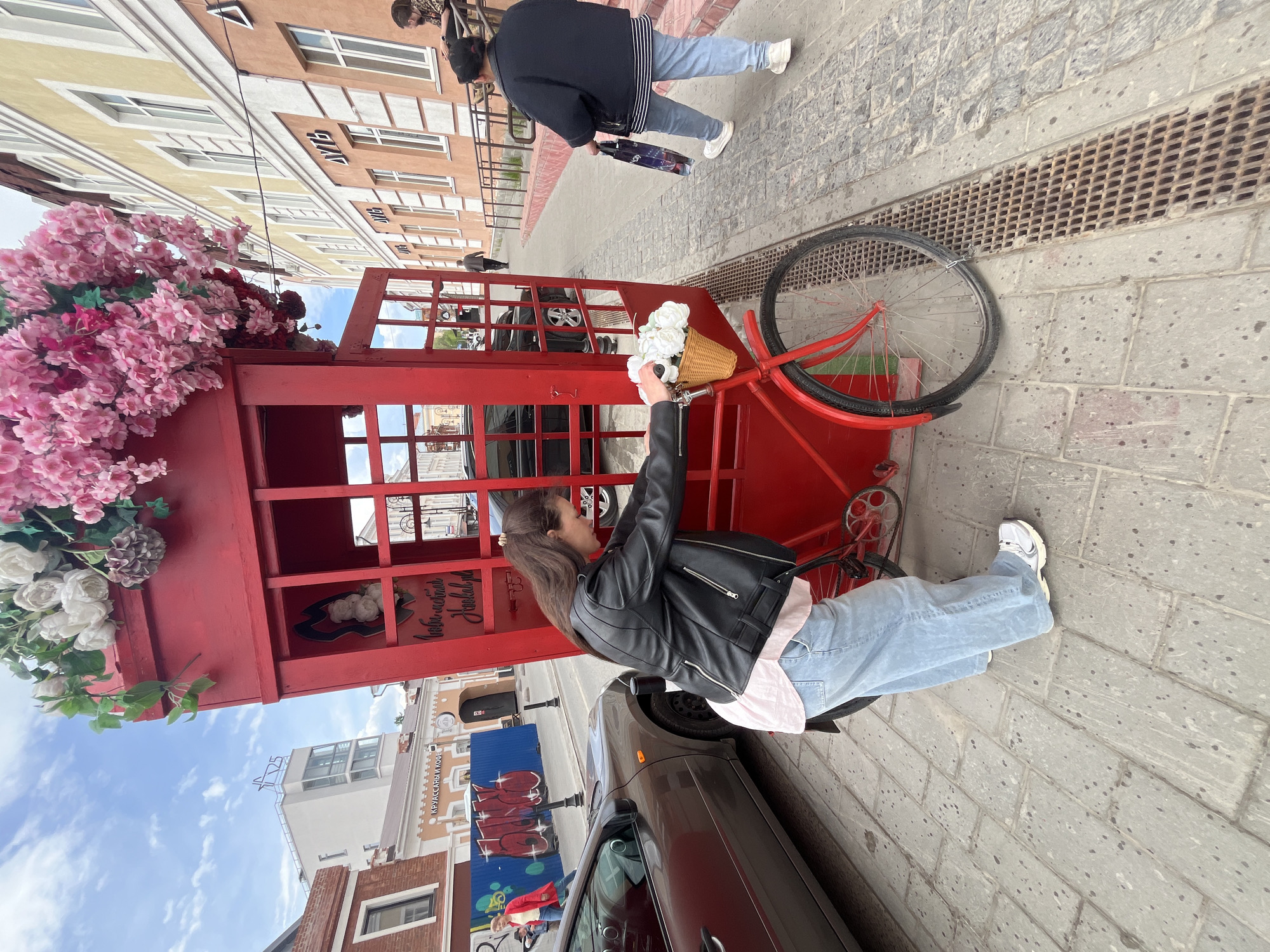 A rotated eye-level photograph shows a red telephone booth decorated with flowers in Ulyanovsk, Russia. A young woman with dark hair wearing a black leather jacket and light blue jeans stands next to the bicycle attached to the phone booth. She is holding white flowers. Buildings line the background with people passing by. A car is parked next to the phone booth. The pavement is gray.