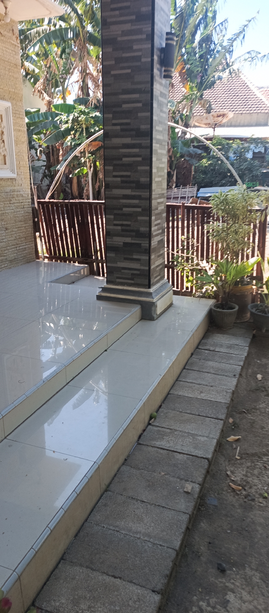 This is a photograph of an outdoor scene in Jatisari, Indonesia, likely a residential area. In the foreground, a tiled pathway with a slight incline leads away from a set of white tiled stairs. A dark, rectangular pillar with a textured, stone-like finish stands prominently to the right of the stairs. Behind the pillar, a wooden fence is visible, with lush green banana trees and other foliage growing beyond it. A house with a brown tiled roof and a satellite dish is partially visible in the background, suggesting a suburban or rural setting. The lighting indicates it is daytime, with sunlight casting shadows. The overall impression is of a well-maintained outdoor space, possibly a porch or garden area.