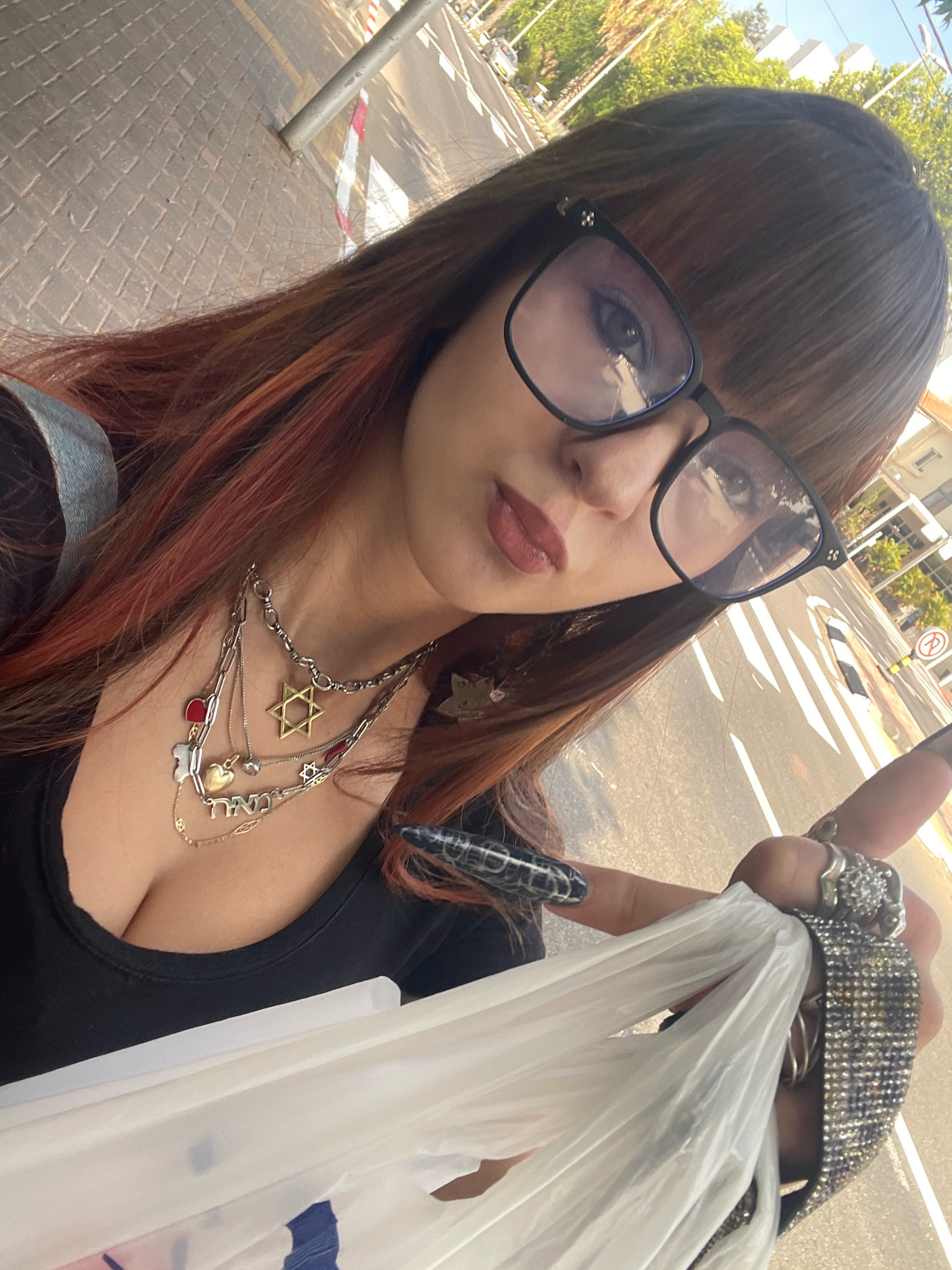 A young woman with auburn-streaked dark hair and bangs, wearing glasses, a black top, and multiple necklaces, is holding a white plastic bag in her right hand. She has elaborate nail art on her fingernails and is wearing several rings on her fingers. The background shows a street scene in Raanana, Israel, with a sidewalk made of paving stones, a street with traffic markings, trees, and buildings. The perspective suggests she is taking a selfie outdoors in an urban environment.