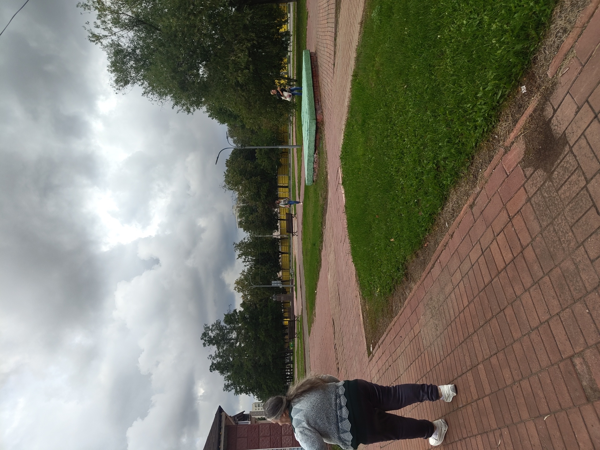 An elderly woman with gray hair pulled back in a ponytail walks on a brick-paved sidewalk in Ramenskoye, Russia. She is wearing a light-colored sweater and dark pants, with white athletic shoes. The scene takes place on a cloudy day, with gray skies overhead. In the background, there are trees, benches, streetlights, and a low green structure. Two other individuals are visible further in the distance; one woman walks past a bench, and another person stands near a green structure holding bags. The environment suggests an outdoor public space like a park or square.