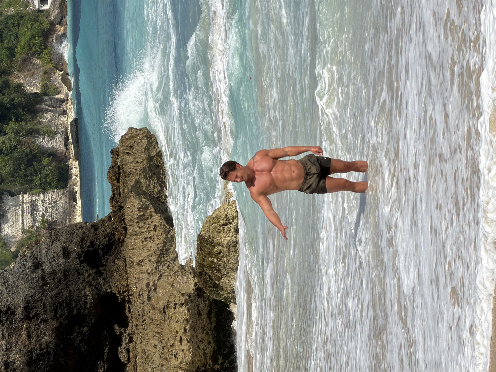 A muscular man in dark swim trunks stands in shallow, foamy ocean water at a beach in Kuta Selatan, Indonesia. He is facing towards the viewer's right, with his right arm extended forward and slightly downward. Behind him, the turquoise ocean churns with waves, and in the background, rocky cliffs covered in lush green vegetation rise up. The sun is shining, casting bright light on the scene.