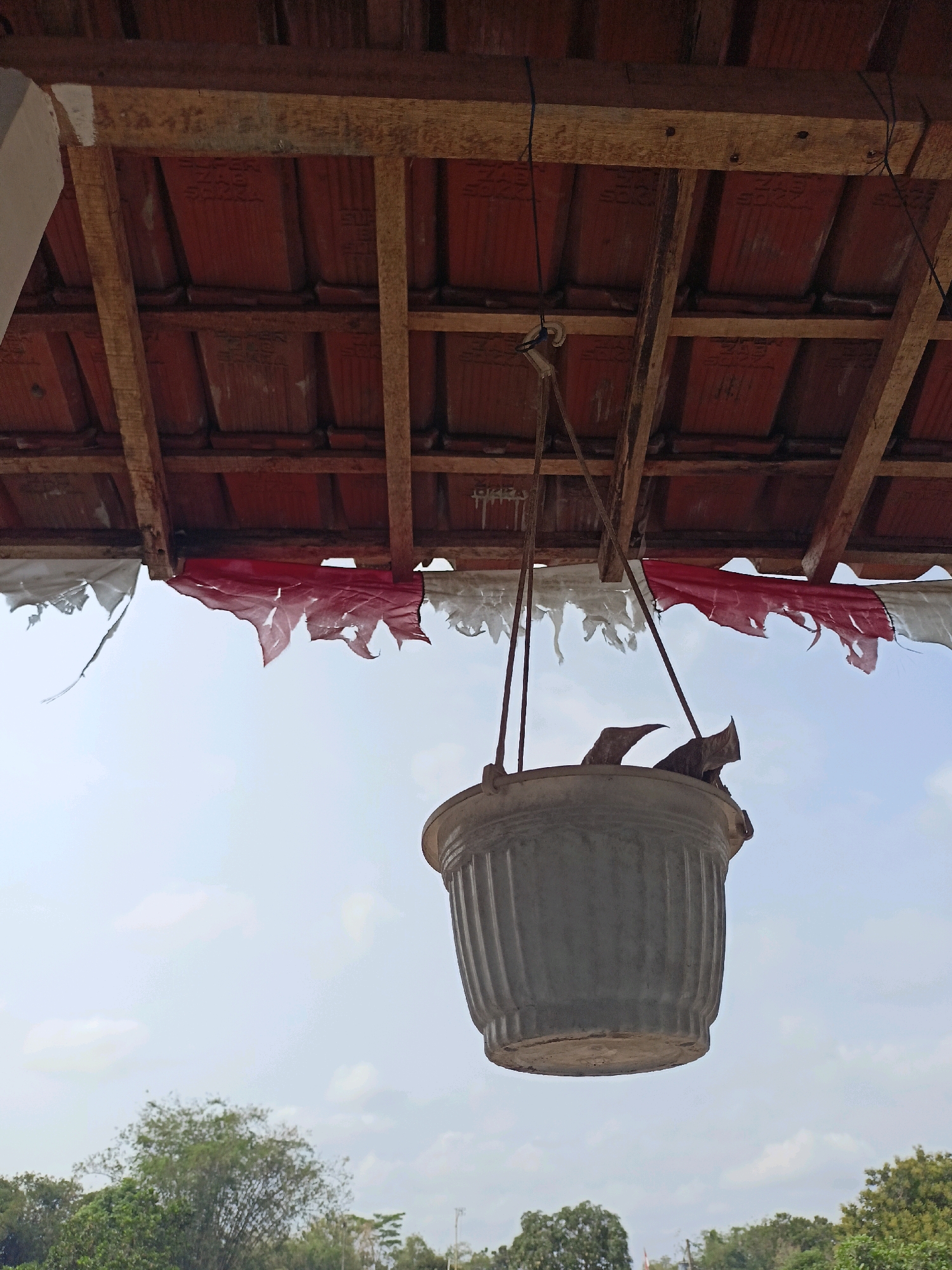The image depicts a grey, ribbed planter hanging from a rustic wooden roof structure. Torn red and white fabric hangs from the underside of the roof, possibly remnants of a flag. The planter appears empty and is suspended by thin wires. The background shows a hazy sky with scattered clouds and a line of green trees and foliage at the bottom of the frame. The scene is set outdoors, likely in Trasan, Indonesia, given the context. There are no people or visible animals in the image. The overall impression is one of a somewhat neglected outdoor space.