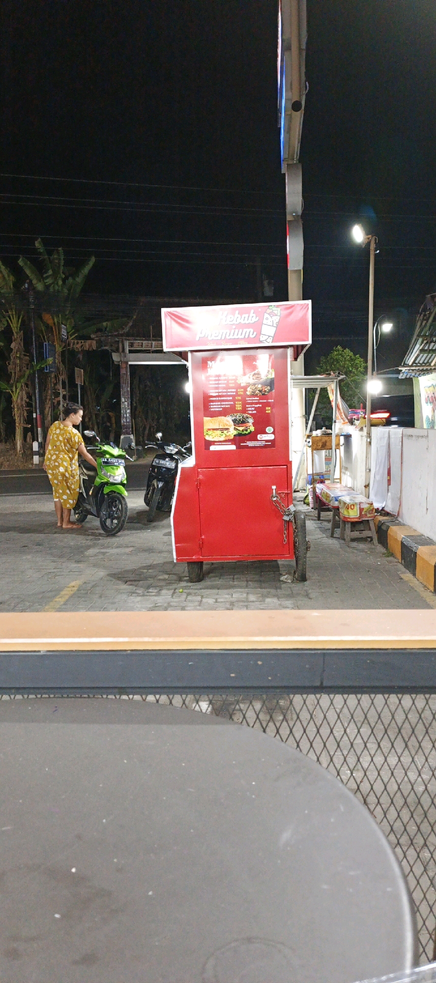 In Godean, Indonesia, at night, a red food cart with a "Kebab Premium" sign is parked on a paved area. A woman in a yellow patterned dress stands next to a green motorcycle. A black motorcycle is parked behind it. The cart has a menu board displaying food items and prices. A chain and lock secure the cart's side door. In the background, there are trees, a building, and a street lamp. The scene appears to be outdoors, possibly a street-side food stall.