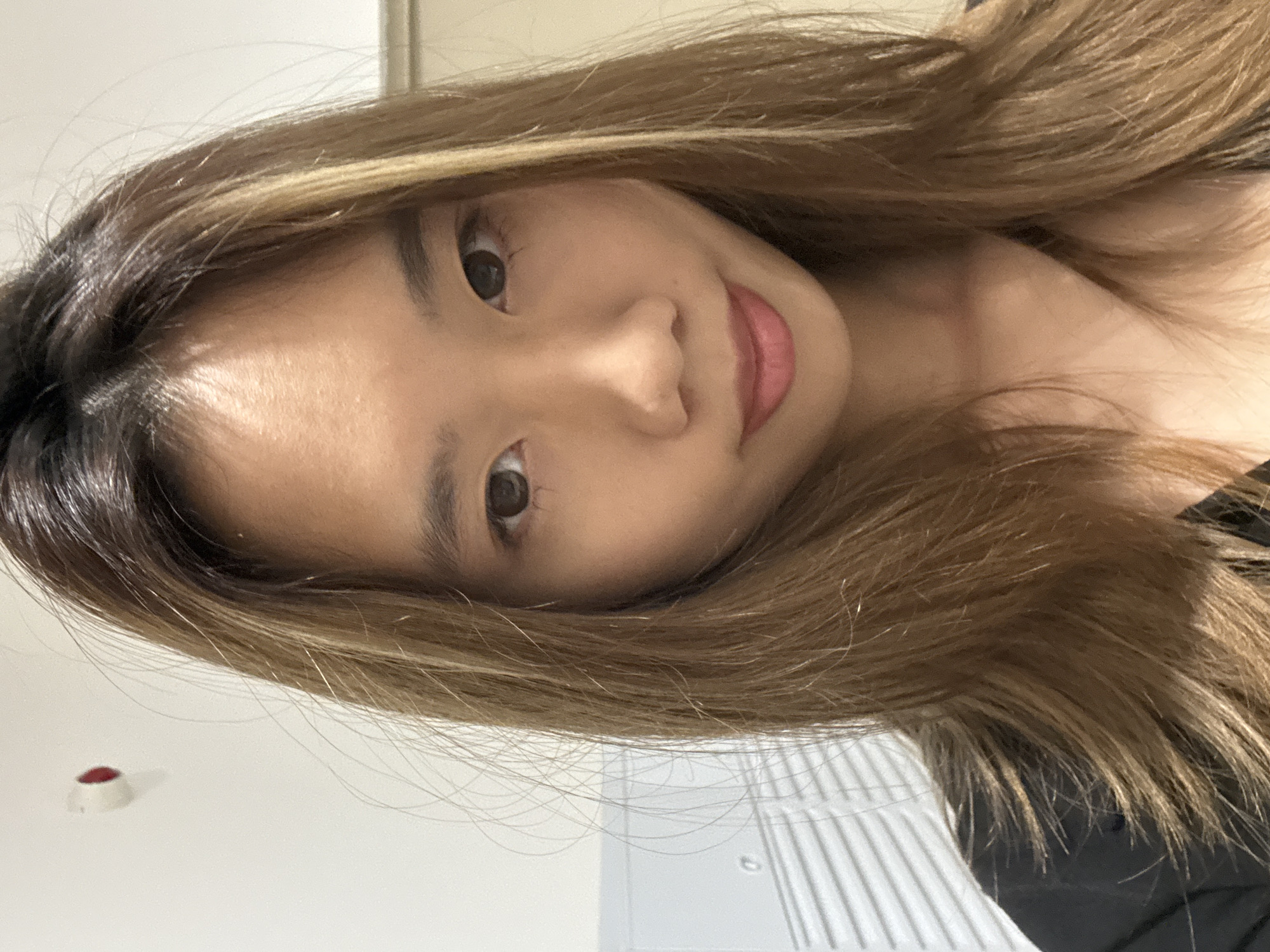 A close-up, selfie-style image of a young woman with long, highlighted brown hair. Her face is slightly angled towards the camera. She has brown eyes, a fair complexion, and is wearing light pink lipstick. The background appears to be a simple interior with white walls and what looks like a window blind. There is a small, round object, possibly a button, visible on the wall behind her. There are no other contextual details to indicate the location in Nan'an District, China.