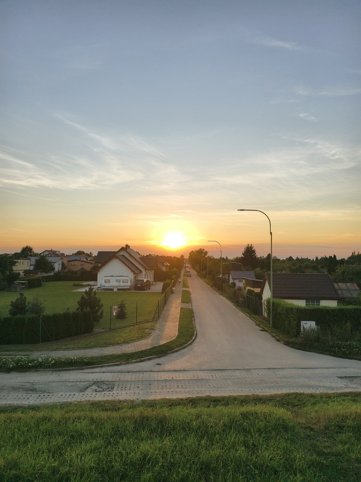 The image shows a quiet suburban street in Tczew, Poland, during sunset. The sun, a bright orb, is setting on the horizon, casting a warm, golden glow across the sky and illuminating the landscape. A paved road stretches into the distance, lined with houses on either side. The houses appear to be residential, with manicured lawns and fences. Some vehicles are visible parked on the properties or on the street. A few people are walking along the sidewalk, enjoying the evening. The sky is a mix of blue and orange, with wispy clouds scattered across it. The overall atmosphere is peaceful and serene.