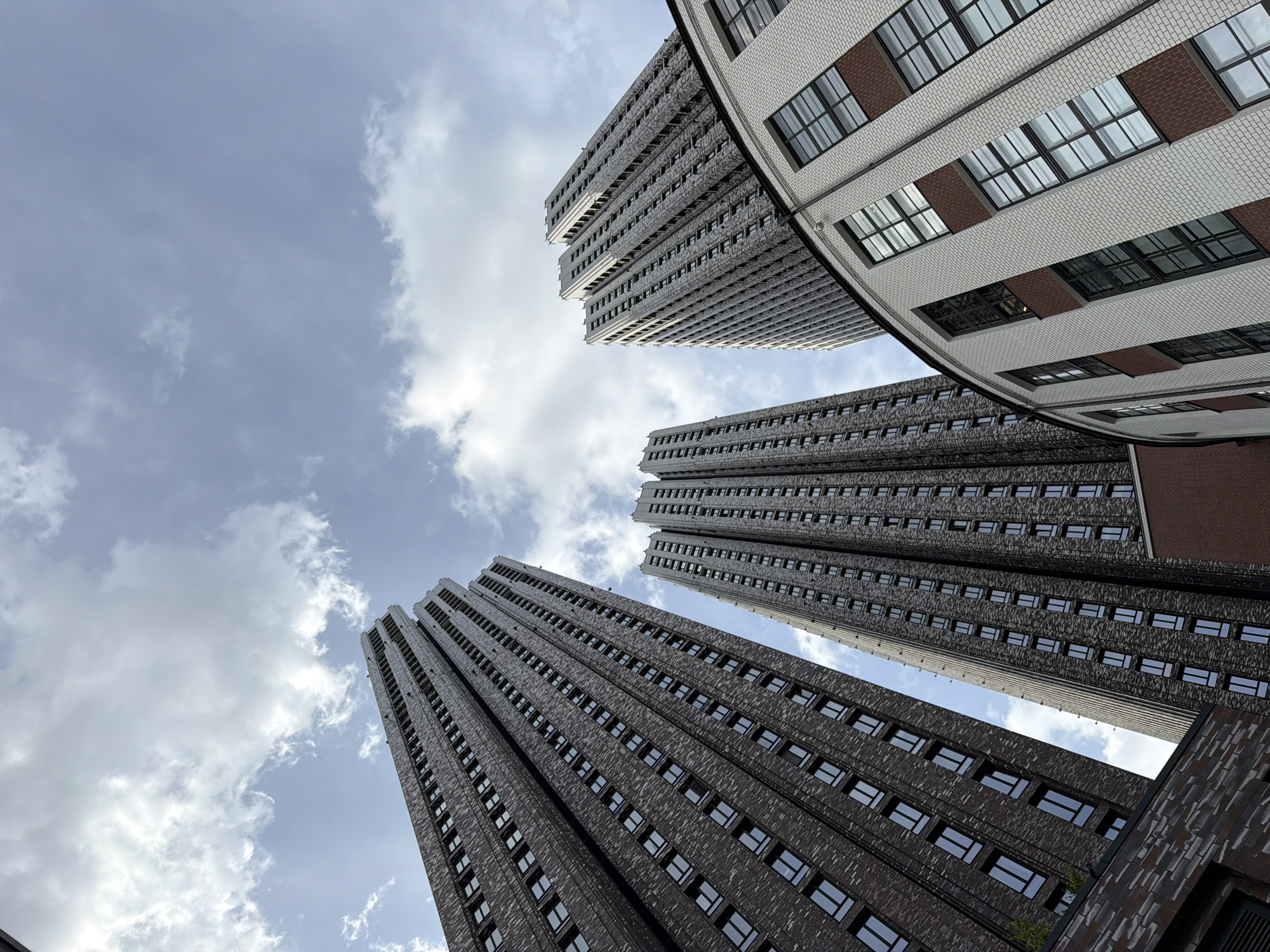The image shows a low-angle view of several tall buildings against a cloudy sky. Three of the buildings are predominantly grey and appear to be of similar design, with a repeating pattern of windows running up their facades. The fourth building is partially visible at the upper right of the frame, showing a curved white brick facade with brown accents and symmetrically spaced windows. The sky consists of a mix of light blue and patches of white clouds.
Based on the architectural style and known landmarks, these buildings appear to be part of the Seven Sisters group of skyscrapers built in Moscow, Russia during the Stalinist era. The buildings in the image are likely located in Moscow. There are no people visible in the image. The focus is on the architecture and the sky.