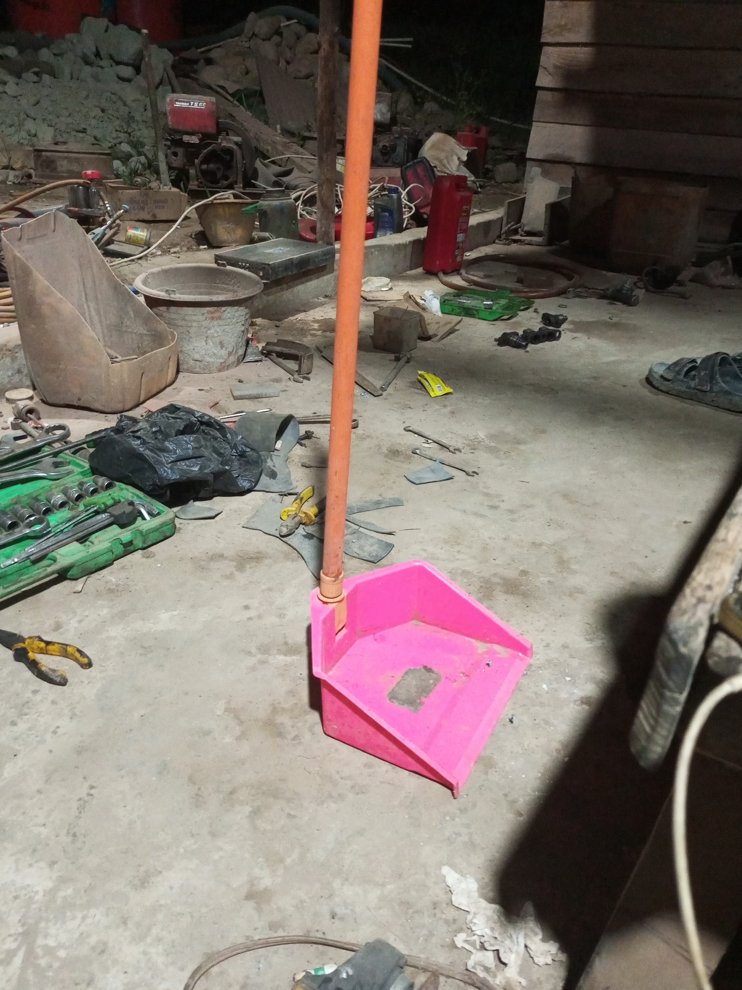 The image depicts a cluttered outdoor scene, possibly a workshop or construction area, in the city of Keling Panggau, Indonesia. The foreground is dominated by a pink dustpan with an orange handle, resting on a concrete surface. Scattered around are various tools, including a green toolbox filled with sockets and wrenches, a pair of yellow-handled pliers, and other assorted hardware. A black garbage bag lies nearby. In the background, construction materials such as piles of dirt and rocks, wooden planks, and what appears to be a small engine or generator are visible. The overall impression is one of disarray, with tools and materials strewn about a utilitarian workspace. The lighting suggests it is either evening or nighttime, with artificial light sources casting shadows. No people are visible in the frame.