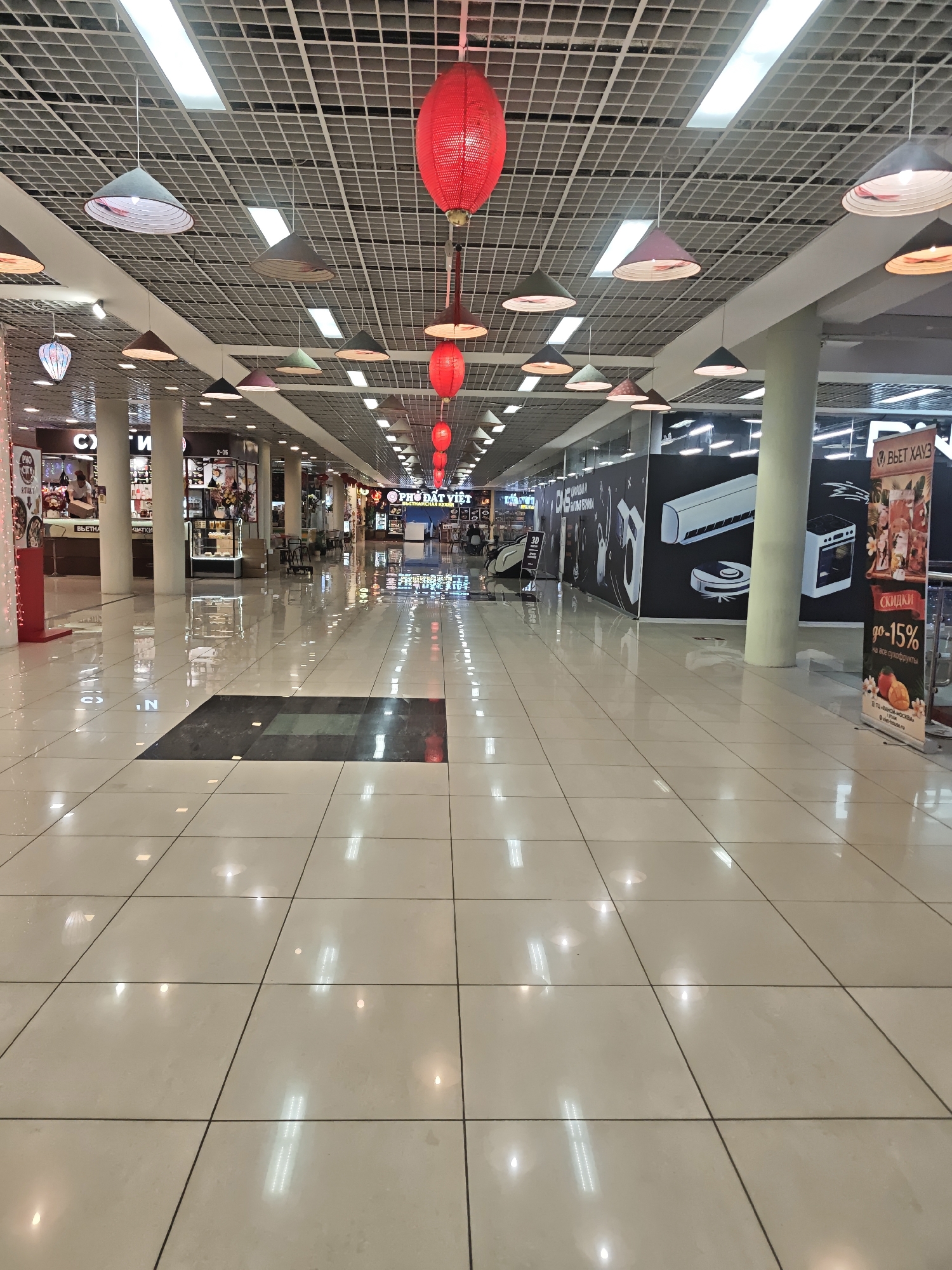 This image captures a long, empty corridor of a shopping center, likely Torgovyi Tsentr "Hanoi-Moscow" (TC "Hanoi Moscow"), given the prominent signage. The setting suggests a commercial space with a distinct Vietnamese cultural theme.
The scene is illuminated by numerous overhead fluorescent lights set into a grid-patterned suspended ceiling. Adding to the ambiance are decorative red, spherical lanterns and conical pendant lights resembling traditional Vietnamese non la hats, hanging at various heights. The floor is made of large, glossy light-colored tiles, highly reflective, showing distorted reflections of the overhead lighting and decorations. A large dark rectangular tile pattern is embedded in the floor further down the corridor.
Along the left side, various shopfronts are visible. "PHO CITY" and "PHỞ ĐẤT VIỆT" signs clearly indicate Vietnamese cuisine establishments. One person is faintly visible behind a counter at "PHO CITY." Other signs like "СУШИ" (Sushi) are also present. On the right, a temporary dark wall is decorated with white outlines of home appliances (an air conditioner, a robot vacuum cleaner, an oven), possibly indicating a future electronics store or a renovation. A display stand advertises "СКИДКИ до -15% на все сухофрукты" (Discounts up to -15% on all dried fruits) from "ВЬЕТ ХАУЗ" (Viet House).
The corridor appears largely deserted, suggesting off-peak hours. The combination of Cyrillic text, Vietnamese shop names, and cultural decorations strongly places the location within a Vietnamese-themed shopping complex in Moscow, Russia.