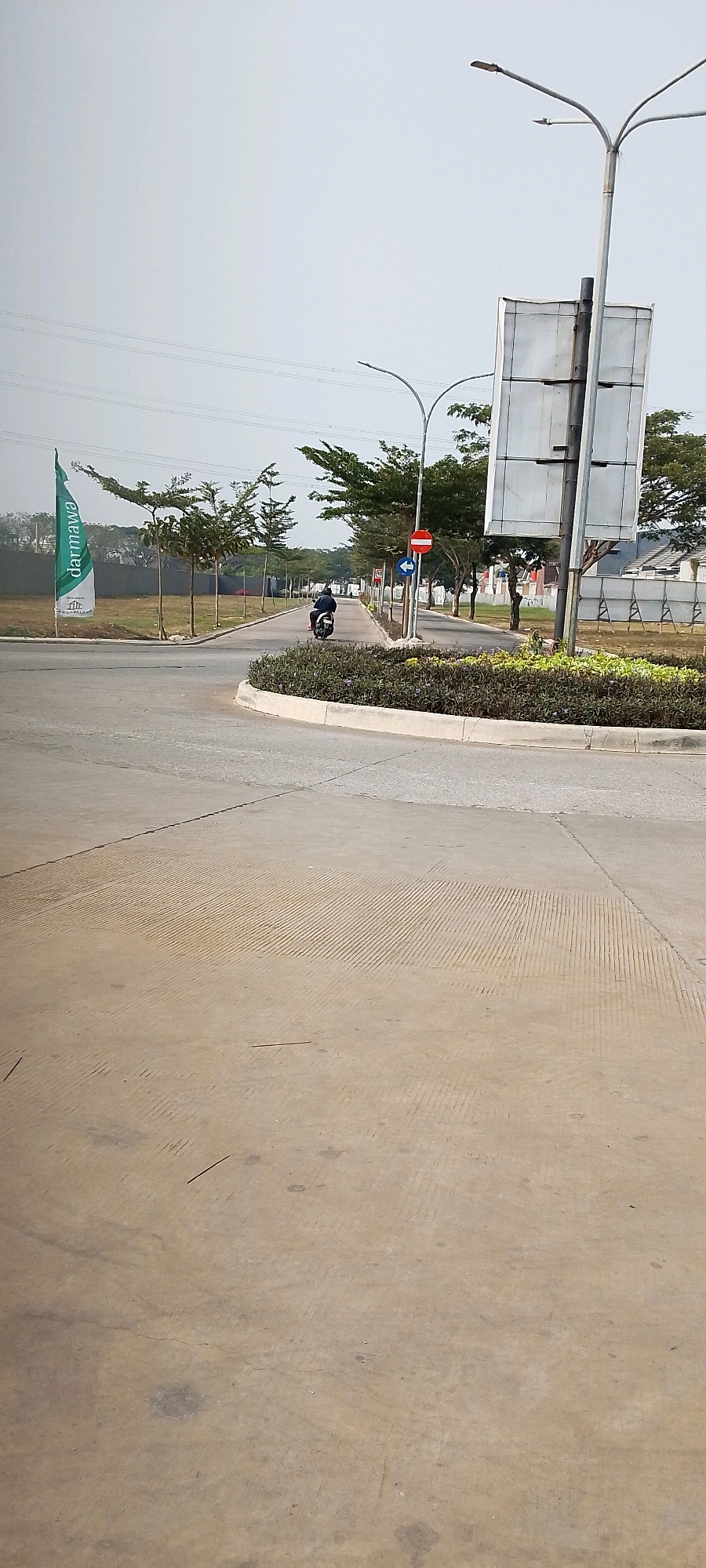 The image depicts a paved road with a motorbike rider heading away from the camera on the right side of the road. The rider is wearing a dark jacket and helmet. The road is bordered by a planted median strip with dark green foliage and concrete curbing. On the left side of the road, a green flag with white lettering that reads "darmawa" is planted in the ground. Behind the flag, there are some buildings and trees. Traffic signs are visible on the right side of the road, including a "no entry" sign and an arrow indicating a left turn. The sky is overcast and the overall environment appears to be a developing area or an industrial park in Satriamekar, Indonesia. The foreground of the image is dominated by a textured concrete surface.