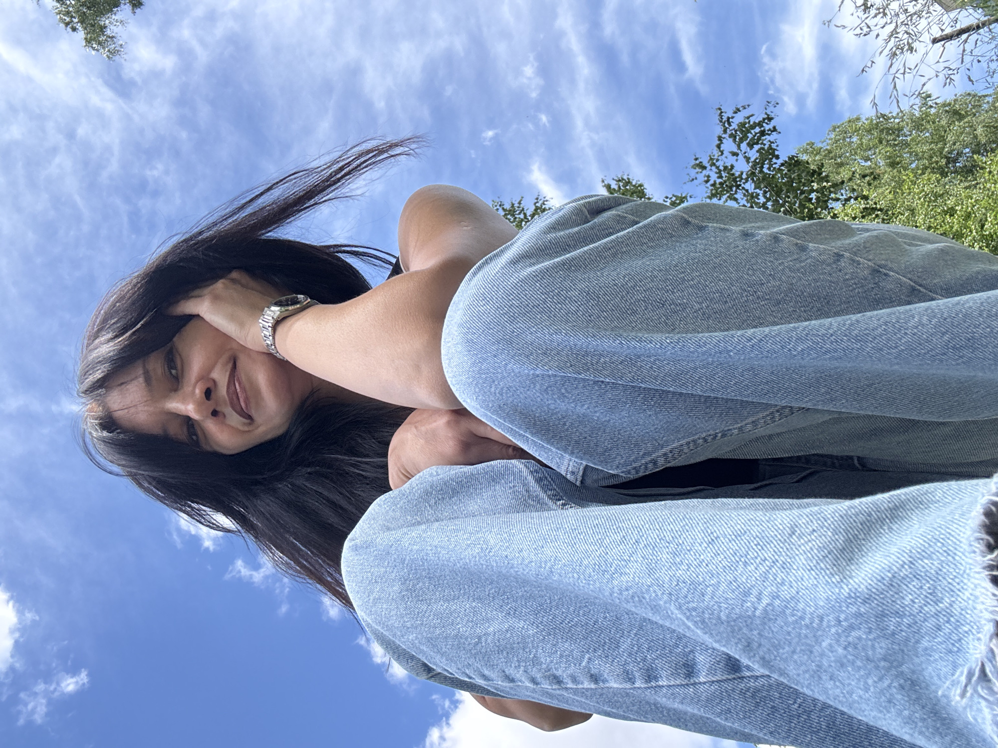A low-angle, close-up shot shows a woman in light-wash denim jeans and a dark top sitting outdoors under a blue sky with wispy clouds. She is facing the camera, with her left arm resting on her bent legs and her right hand touching her face. Her long, dark hair is blowing gently. A silver wristwatch is visible on her right wrist. In the background, green trees are visible against the sky. The image appears to have been taken in Chaikovsky, Russia, during the daytime.