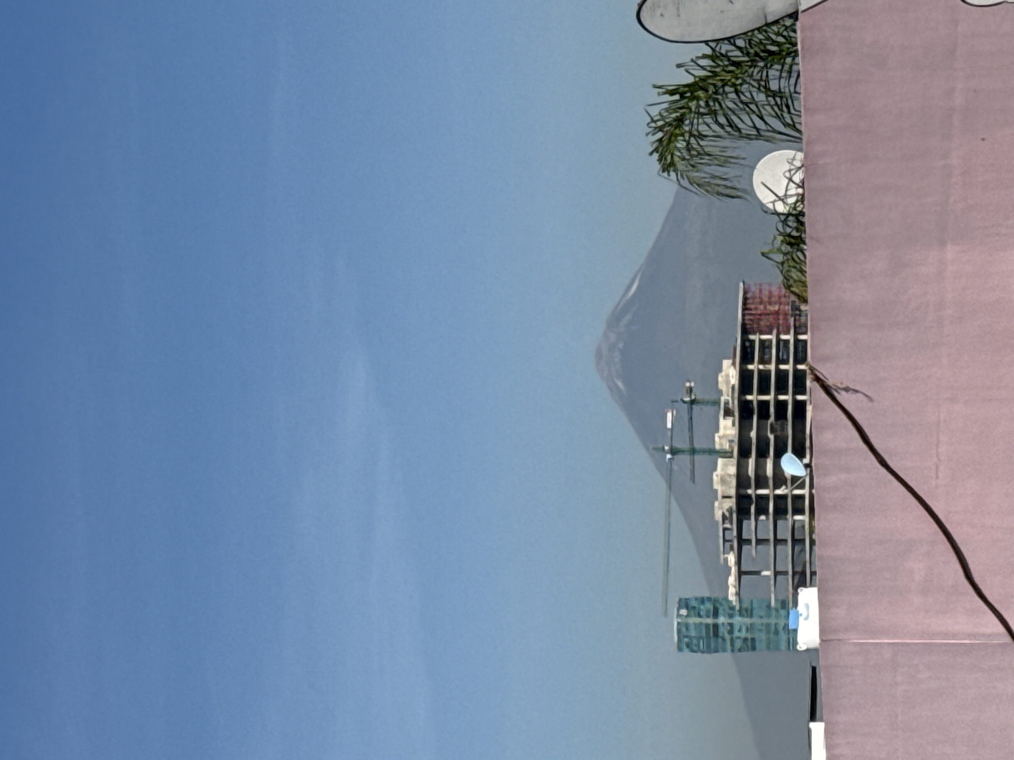 The image shows a view in Puebla, Mexico, with a clear blue sky dominating the left side. A mountain is visible in the distance, partly obscured by haze or cloud cover. In the foreground, there's a building under construction, with exposed framework and visible construction cranes. To the right, a pink wall-like structure partially obscures the building. There are trees present on the top of the building.