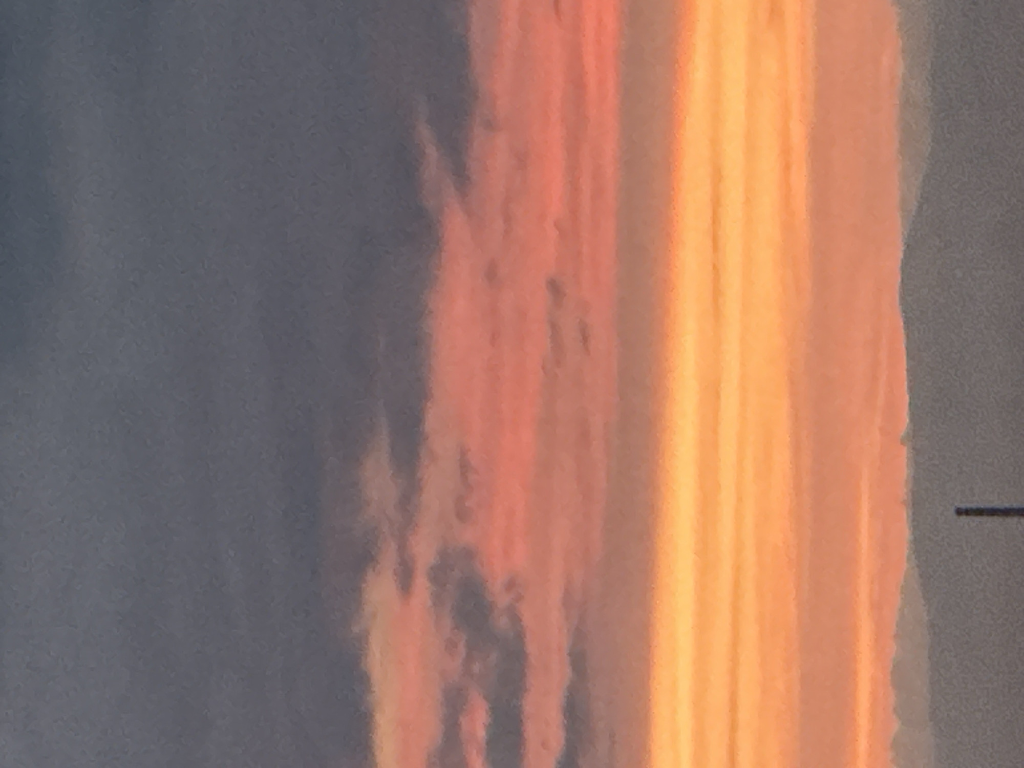 The image shows a cloudy sky during sunset or sunrise, with bands of grey, pink, and orange hues. The lower portion of the image depicts a silhouetted mountain range, with a faint industrial structure, possibly a chimney, visible against the sky in the foreground. The scene is devoid of any human presence or activity. The location is specified as Asenovgrad, Bulgaria.