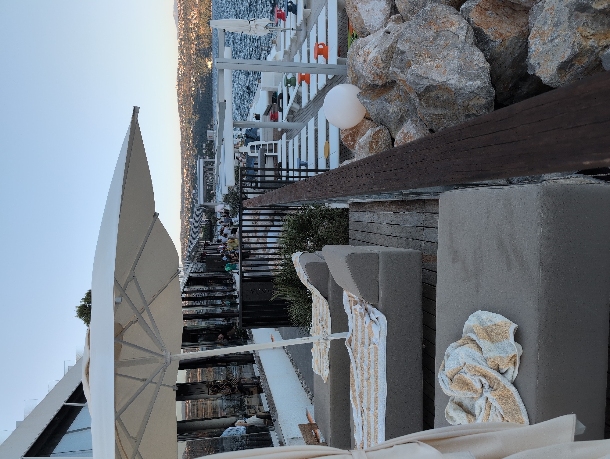 The image depicts an outdoor seating area at what appears to be a resort or restaurant, situated in the city of Opatija, Croatia, along the Adriatic Sea. In the foreground, two empty lounge chairs are set up on a wooden deck, each draped with a striped towel. A large white umbrella provides shade over one of the chairs. Beyond the chairs, the scene opens up to reveal a view of the sea and a distant, populated coastline with hills.
To the left, a modern building with large windows, possibly part of the resort or restaurant, is visible. Inside, several people are seen, suggesting indoor activity. In the mid-ground, rows of white lounge chairs and tables with colorful stools are arranged, implying a communal relaxation or dining area. Some individuals are present in this area, engaged in various activities.
The environment is characterized by the juxtaposition of a man-made structure and a natural coastal landscape. The sea is calm, reflecting the clear sky. The shoreline is densely covered with vegetation and buildings, indicating a developed urban area. The overall impression is that of a leisure and hospitality setting, likely a beach club or waterfront restaurant, enjoying the scenic beauty of Opatija.