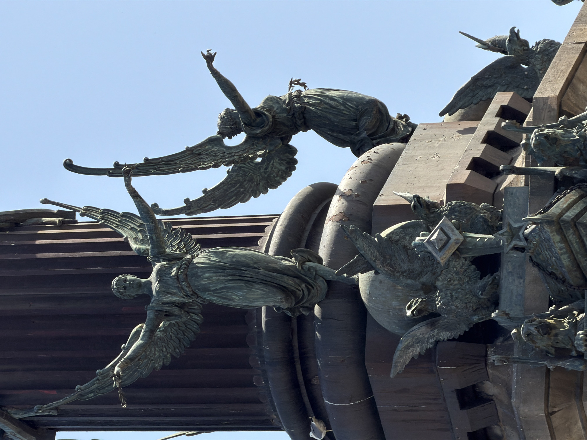 This media file features a close-up, low-angle shot of a bronze monument against a clear blue sky, suggesting it is daytime with fair weather. The monument depicts several figures that appear to be angels or winged beings, rendered in a dark, weathered bronze. The composition focuses on the intricate details of the sculptures, highlighting their dynamic poses and draped clothing.
In the foreground, a winged figure is seen in mid-flight or falling, with its body arched and arms outstretched. Its wings are spread, showing detailed feathering, and its head is turned downwards. Behind this figure, another angel is depicted in a kneeling or prostrate position, with one arm raised towards the sky and wings also prominently displayed. This figure appears to be reaching for something or in a state of supplication. Further to the right and partially obscured, other sculptural elements suggest a larger, complex monument with more winged figures and possibly architectural components. These include what look like layered structures, possibly stairs or platforms, and other sculpted figures with bird-like features or elaborate ornamentation. The overall impression is one of dramatic movement and possibly an allegorical or commemorative scene. The weathered patina of the bronze suggests age and exposure to the elements, hinting at the monument's historical significance. The clarity of the sky and the bright sunlight cast distinct shadows on the sculptures, emphasizing their three-dimensional form. Although no text is explicitly visible, the style of the sculpture could suggest a historical or religious context, possibly a war memorial or a monument to victory, given the heroic and dramatic representations of the winged figures. Given the provided location context of Barcelona, Spain, this could be a part of a well-known monument in the city.