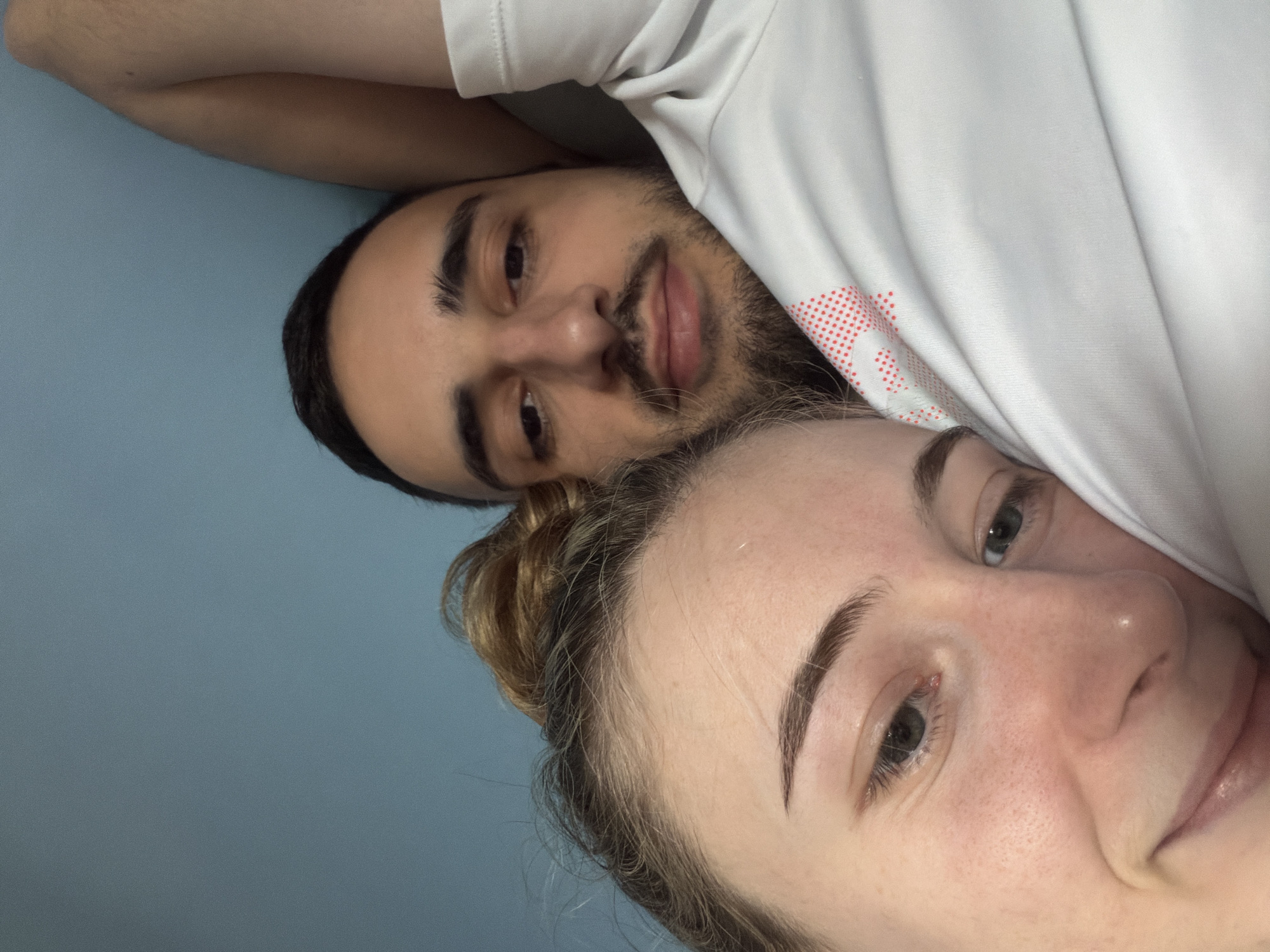 A close-up, tilted, indoor shot captures two individuals. A man with dark hair, a beard, and a white T-shirt with a small red dotted design is at the top. A woman with her hair in a bun is at the bottom. They are likely lying down. The background is a solid light blue. The image does not provide information about the location in Plovdiv, Bulgaria.
