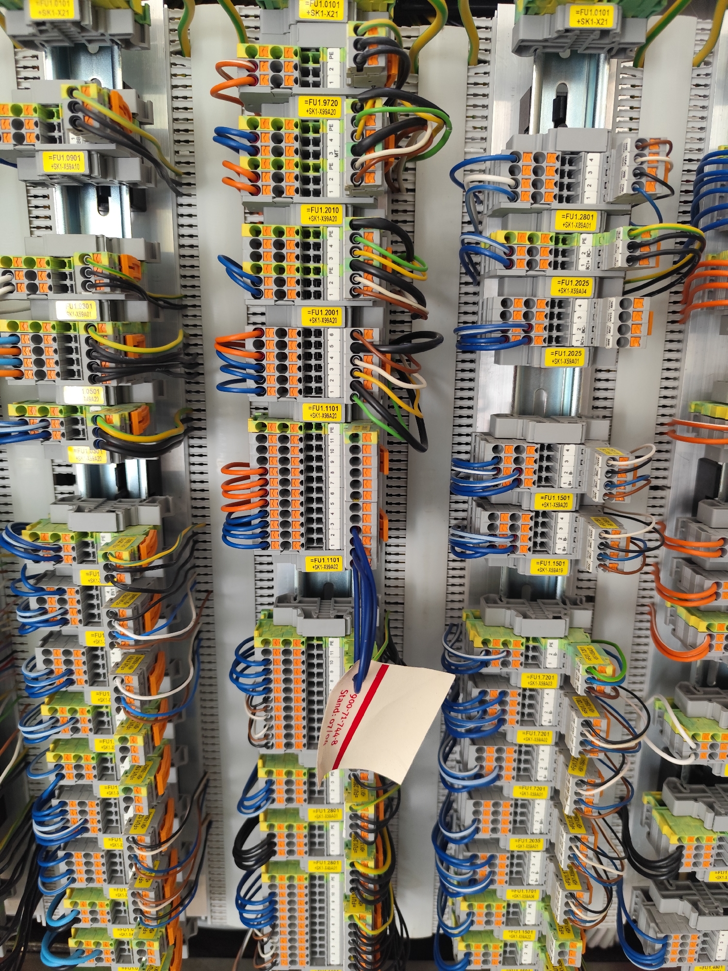 The image shows a close-up of an industrial electrical panel with numerous wires, connectors, and terminal blocks. The panel is mounted vertically. The terminal blocks are gray and orange, with yellow labels indicating component identifiers such as "=FU1.0101 +SK1-X21" and "=FU1.9720 +SK1-X99A20". Various colored wires, including blue, orange, yellow, green, black, and white, are connected to these terminals. Some wires are neatly coiled, while others are bundled together. A white rectangular tag with red writing is partially visible on the right side of the panel. The environment appears to be an indoor electrical or automation control room. There is no information in the image to suggest a specific location in Teba, Indonesia.