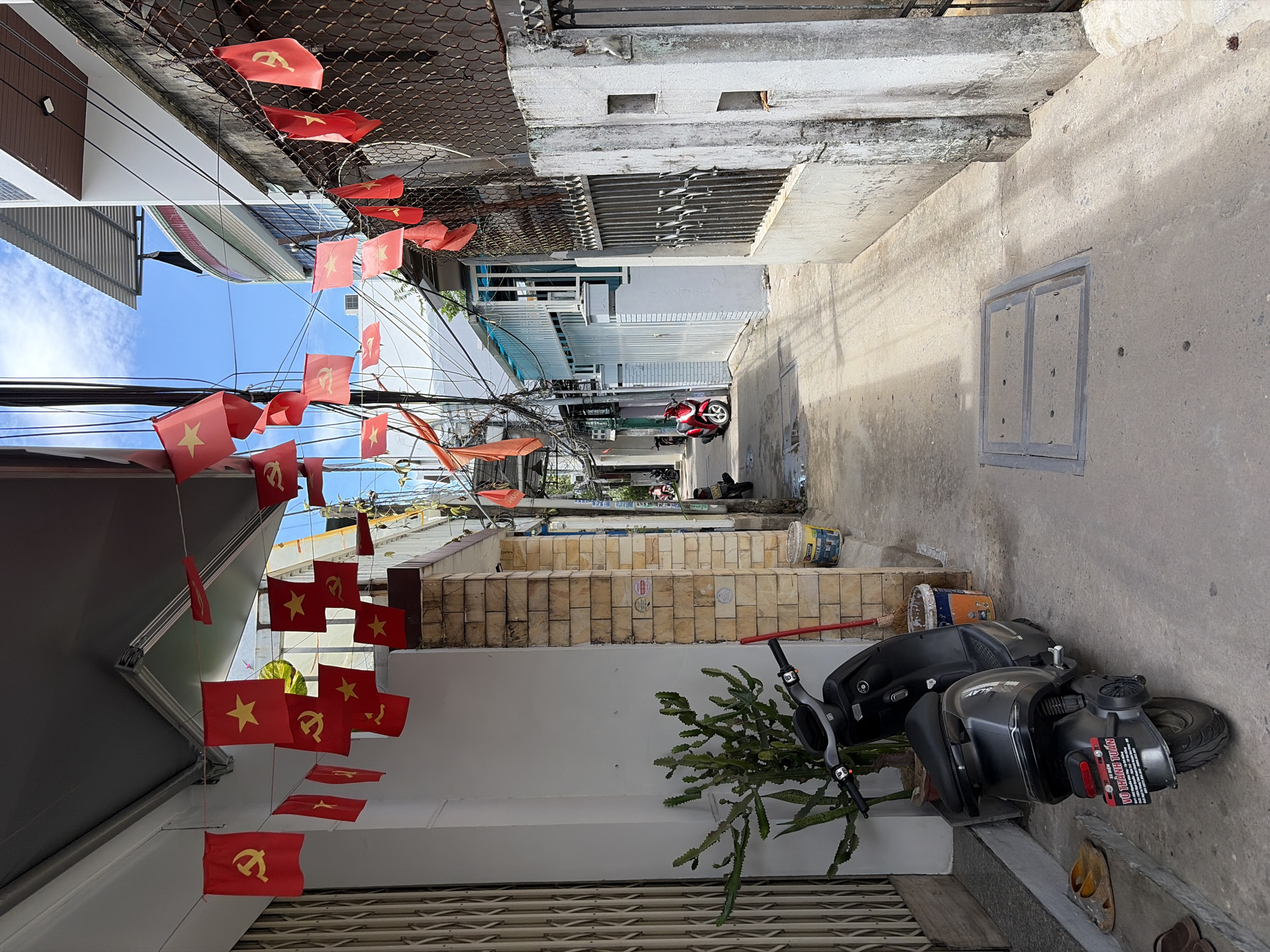 The image depicts a narrow alleyway in Da Nang, Vietnam, decorated with numerous red and yellow Vietnamese flags. The flags are hung on strings across the alley, some with the yellow star and hammer and sickle symbol, indicative of the Vietnamese Communist Party. A dark gray scooter is parked on the left side of the alley, in front of a white wall and a potted cactus plant. Further down the alley, another red scooter is visible, along with a fence and gate on the right side. The alley has a concrete ground, with some puddles of water, and overhead wires. Buildings line both sides of the alley, some with balconies and corrugated metal roofs. The sky is blue with scattered clouds. The scene appears to be an everyday street view in a residential or commercial area of the city.