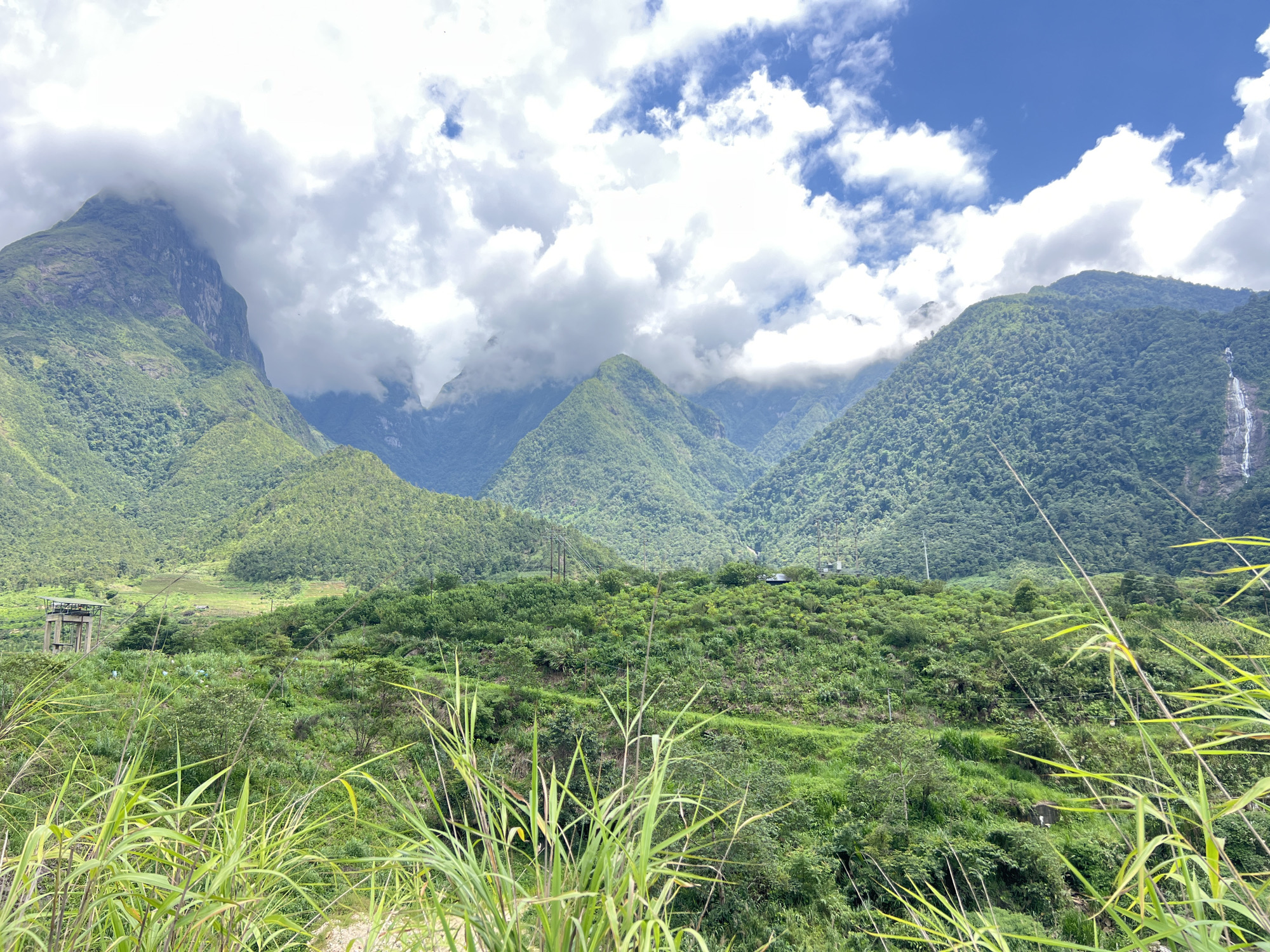 This is a wide shot of a lush green mountainous landscape in Bình Lư, Vietnam. The foreground is dominated by tall green grasses in sharp focus, with a blurred background of rolling hills covered in dense vegetation. In the mid-ground, a vast expanse of green slopes upward, dotted with trees and some signs of human activity. To the left, a concrete structure resembling a water tower or industrial facility is visible. Further into the background, a series of mountain peaks rise, partially shrouded by dramatic white clouds against a bright blue sky. On the far right of the image, a thin waterfall cascades down the side of one of the mountains. The overall impression is one of natural beauty and rugged terrain.