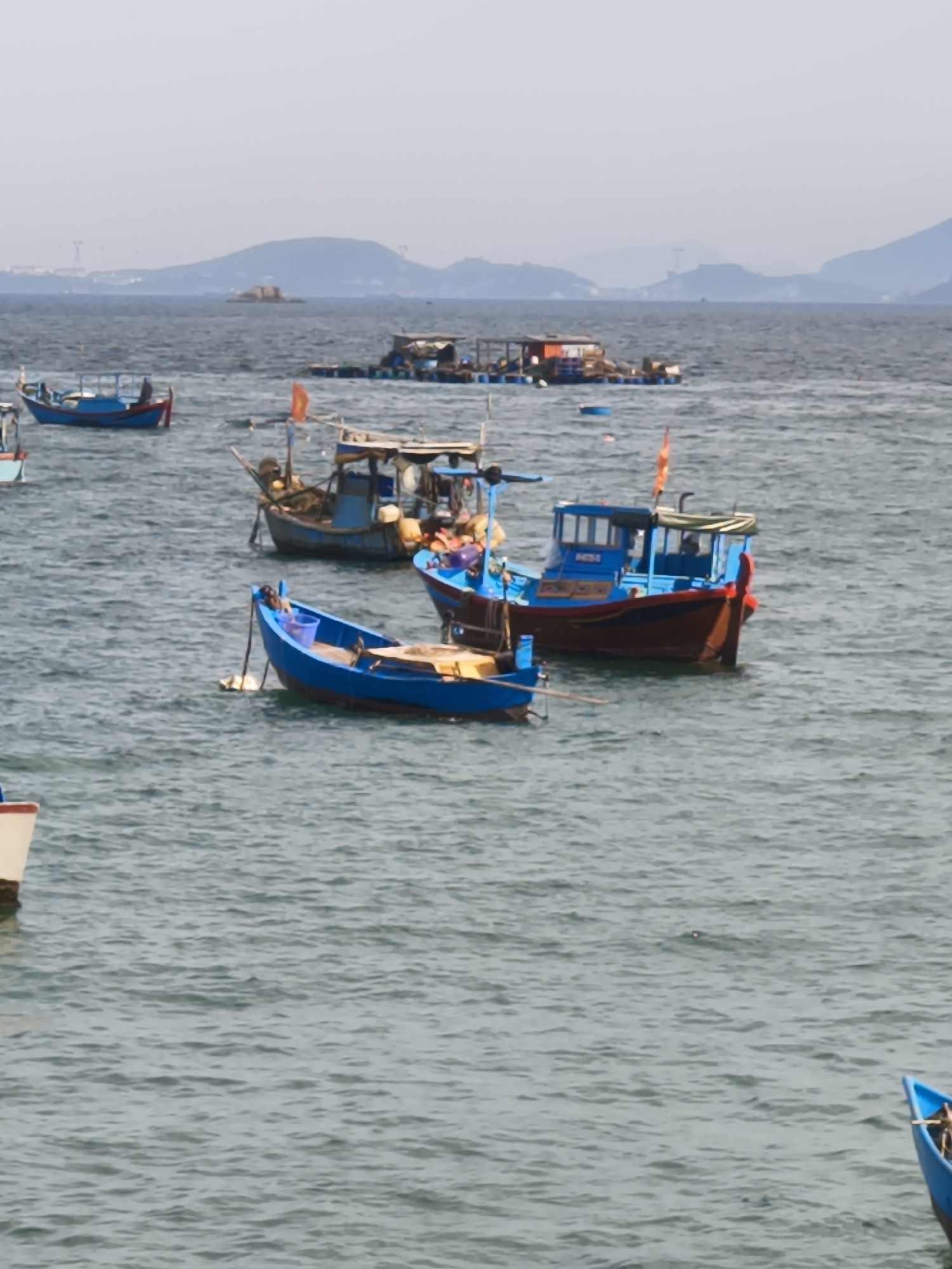 The image depicts a serene scene of various fishing boats anchored in the calm waters of Nha Trang, Vietnam. In the foreground, a bright blue fishing boat is prominently displayed, its hull gleaming under the overcast sky. To the left, another boat with a white and beige hull is partially visible, hinting at the presence of more vessels further out.
In the mid-ground, several other fishing boats are scattered across the water. A blue and red fishing boat, larger than the ones in the foreground, stands out with its multi-level structure and a green canopy. A smaller, darker blue boat with a similar design is positioned behind it, suggesting a fleet of working vessels. A floating structure, possibly a raft or a small house, is visible in the background, adding to the sense of a working harbor or fishing village.
The background is dominated by a hazy, mountainous landscape that extends across the horizon, characteristic of Nha Trang's coastal geography. The water is a muted gray-blue, with subtle ripples indicating a gentle movement. The overall atmosphere is tranquil, with a sense of quiet activity typical of a fishing community. There are no people clearly visible in the image, but the presence of multiple boats suggests human activity related to fishing. The scene is a realistic portrayal of a coastal area in Nha Trang, known for its vibrant fishing industry.