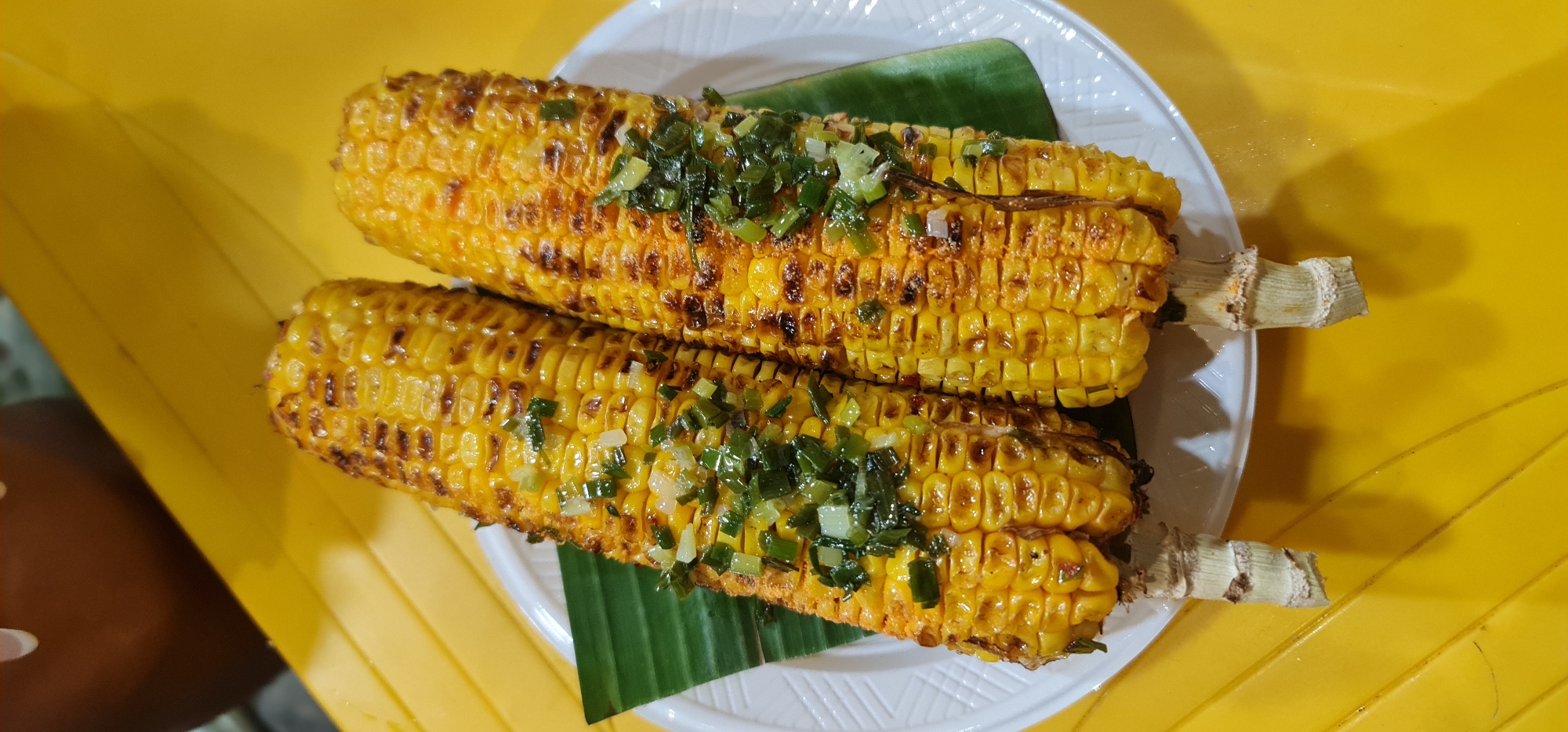 Two grilled corn on the cobs are placed on a white plate over a banana leaf. The corn is topped with chopped green onions. The plate sits on a yellow surface. The scene is likely in Đà Nẵng, Vietnam. No people are fully visible.