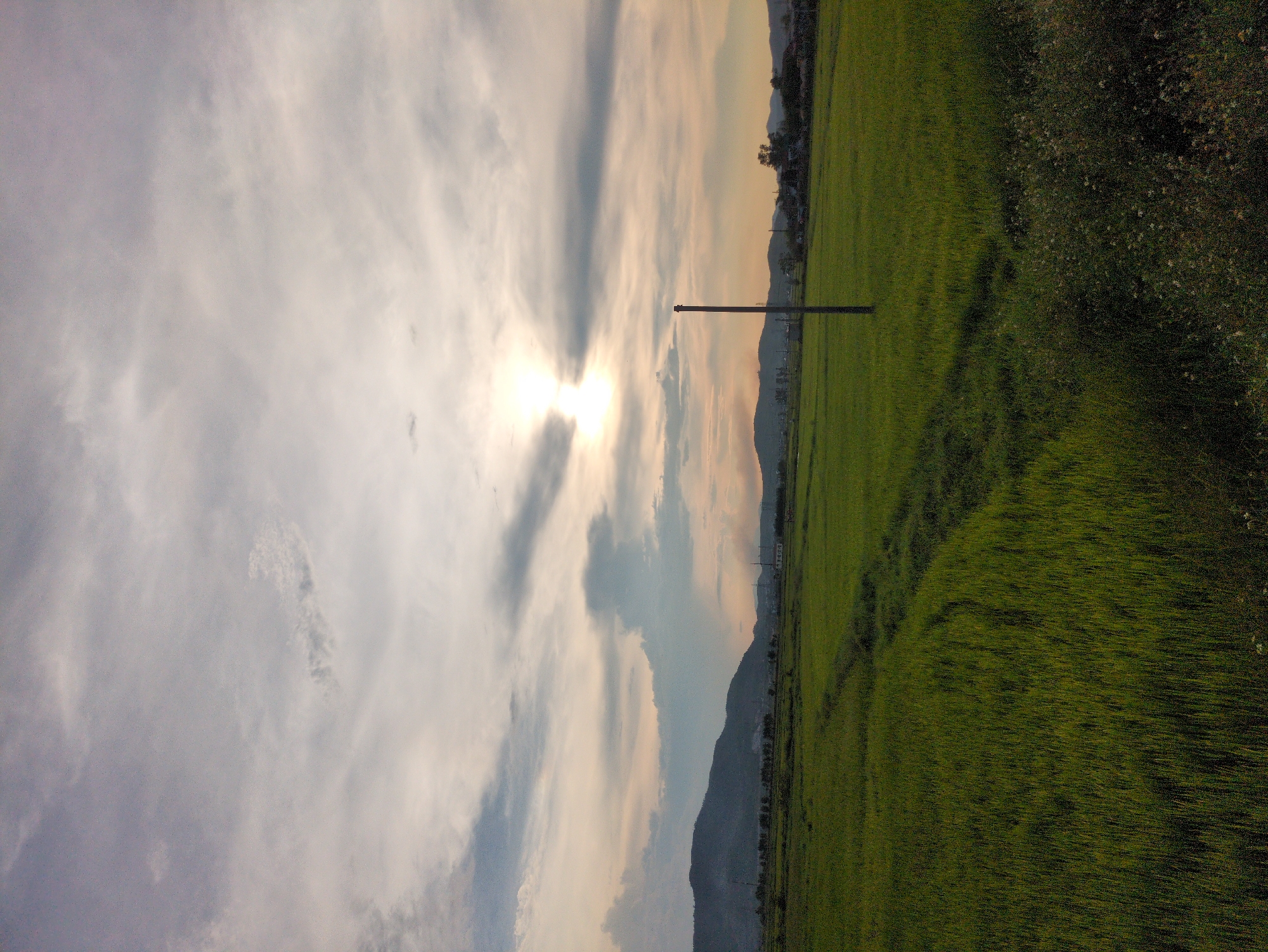 This is a photograph of a rural landscape in Kỳ Khang Commune, Vietnam, during what appears to be either sunrise or sunset. The foreground is dominated by a vibrant green rice paddy field, with subtle tire tracks indicating recent agricultural activity. A single wooden post stands prominently in the middle ground, breaking the expanse of the field. Beyond the field, a line of trees and low-lying buildings mark the edge of a settlement, with rolling hills and mountains forming the backdrop. The sky is filled with a mix of clouds, with the sun peeking through, casting a soft, diffused light over the scene. The overall impression is one of tranquility and natural beauty.