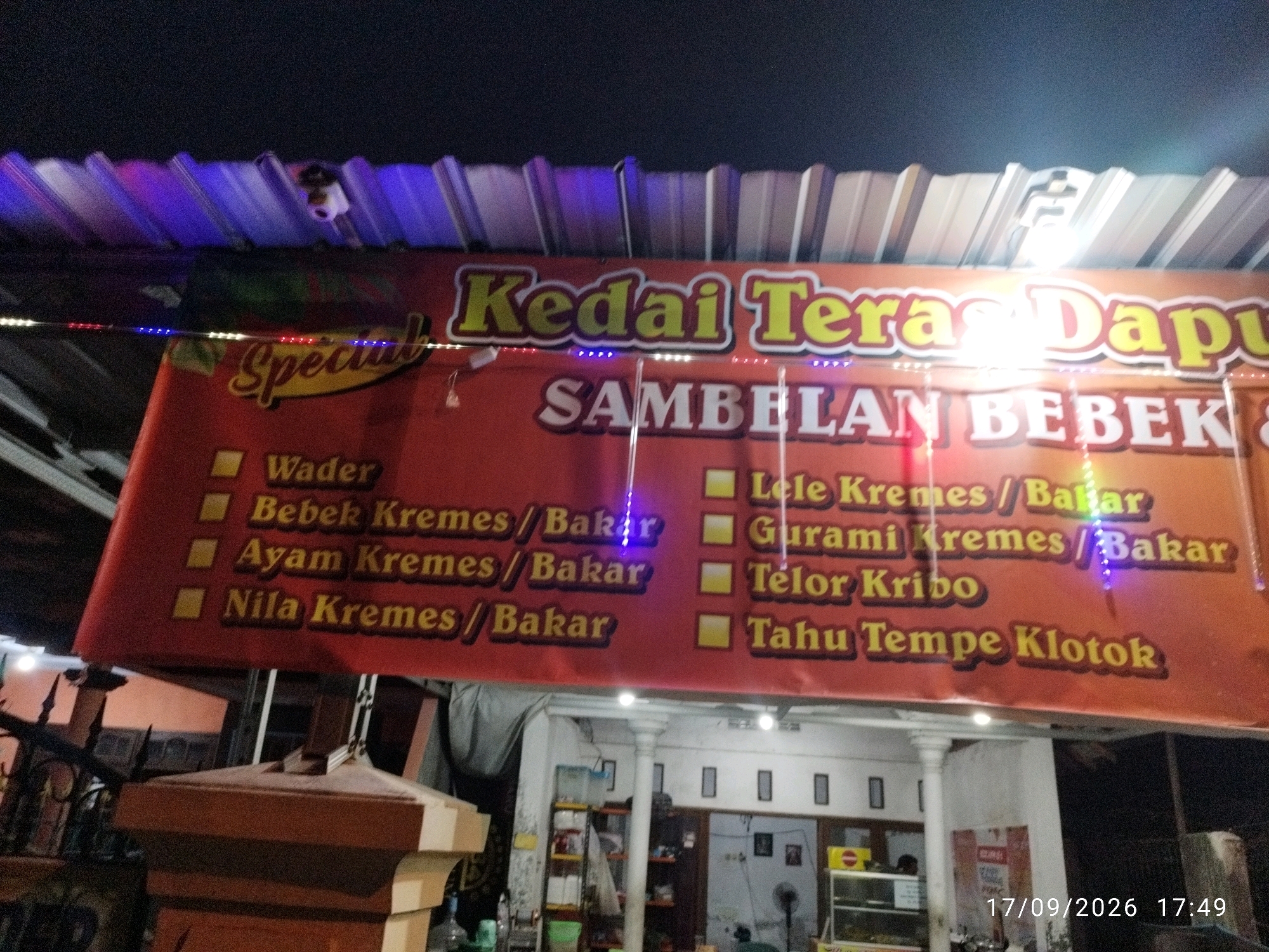 The image shows a food stall sign in Mojokerto, Indonesia. The sign is red with yellow lettering and lists various dishes including "Wader," "Bebek Kremes/Bakar," "Ayam Kremes/Bakar," "Nila Kremes/Bakar," "Lele Kremes/Bakar," "Gurami Kremes/Bakar," "Telor Kribo," and "Tahu Tempe Klotok." The stall appears to be located in front of a building with a white pillar and a corrugated metal roof. The date and time stamp on the image indicate it was taken on September 17, 2026, at 17:49. The overall scene suggests a local eatery operating in an urban or semi-urban area.