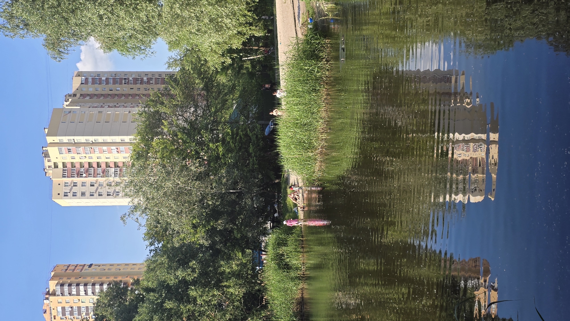 This is a vertical, eye-level shot of a reflective body of water in Kyiv, Ukraine, with a sandy shore and a backdrop of trees and apartment buildings. The water occupies the lower half of the frame, reflecting the sky, trees, and buildings. On the shore, a group of people are present, including children playing near the water's edge and adults relaxing. The shore is lined with tall green grass and trees. In the distance, two multi-story apartment buildings with light-colored facades and numerous windows are visible against a clear blue sky. The scene appears to be a leisure spot within the city.