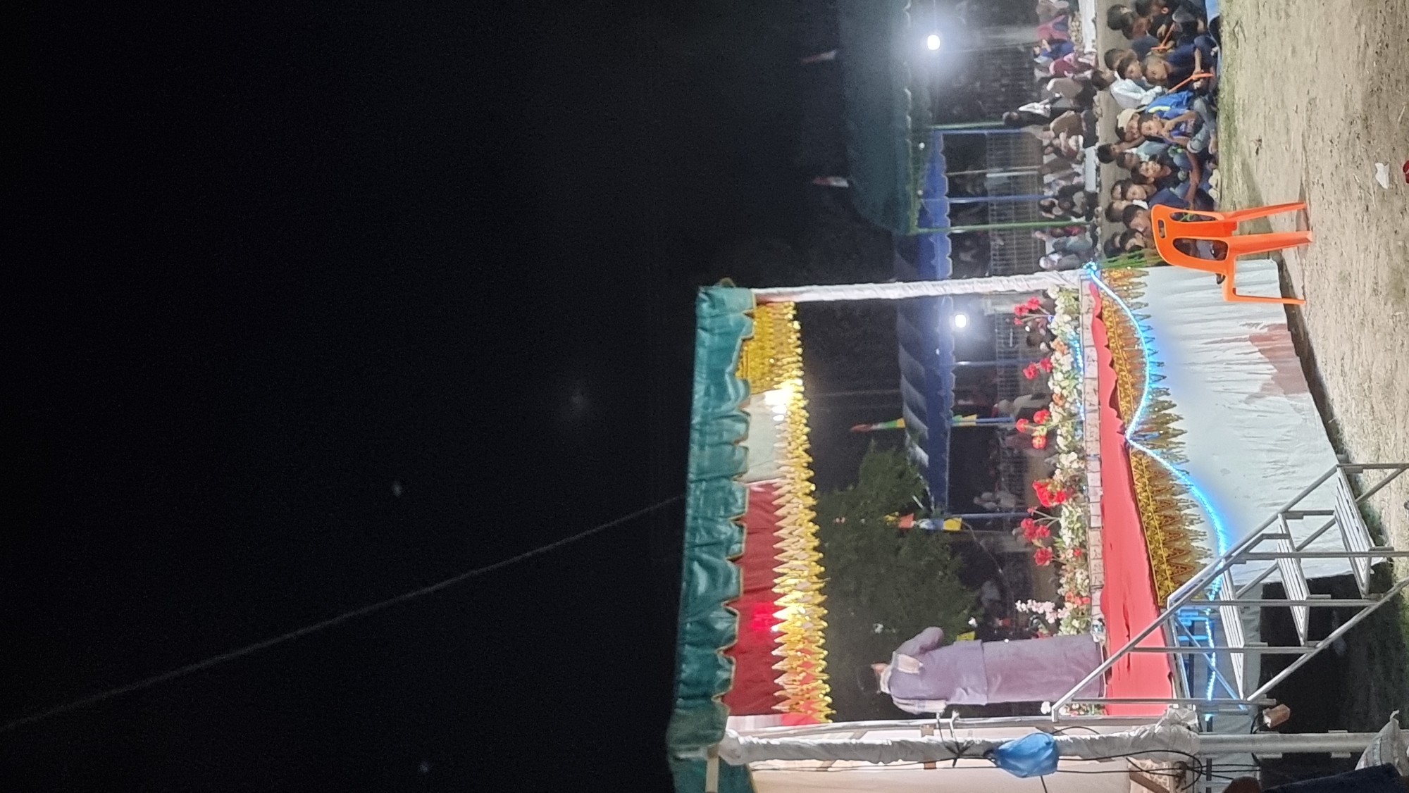 At night in Tumpokbeurandang, Indonesia, a stage is set up for an event. The stage is decorated with a red carpet, golden trim, and blue string lights. A person in a purple outfit stands on the stage facing away from the camera. In front of the stage, an orange plastic chair sits on the ground. To the right, a crowd of people, mostly children, are seated on the ground, facing the stage. Tents are visible in the background, along with more people gathered. The sky is dark and appears to be clear.