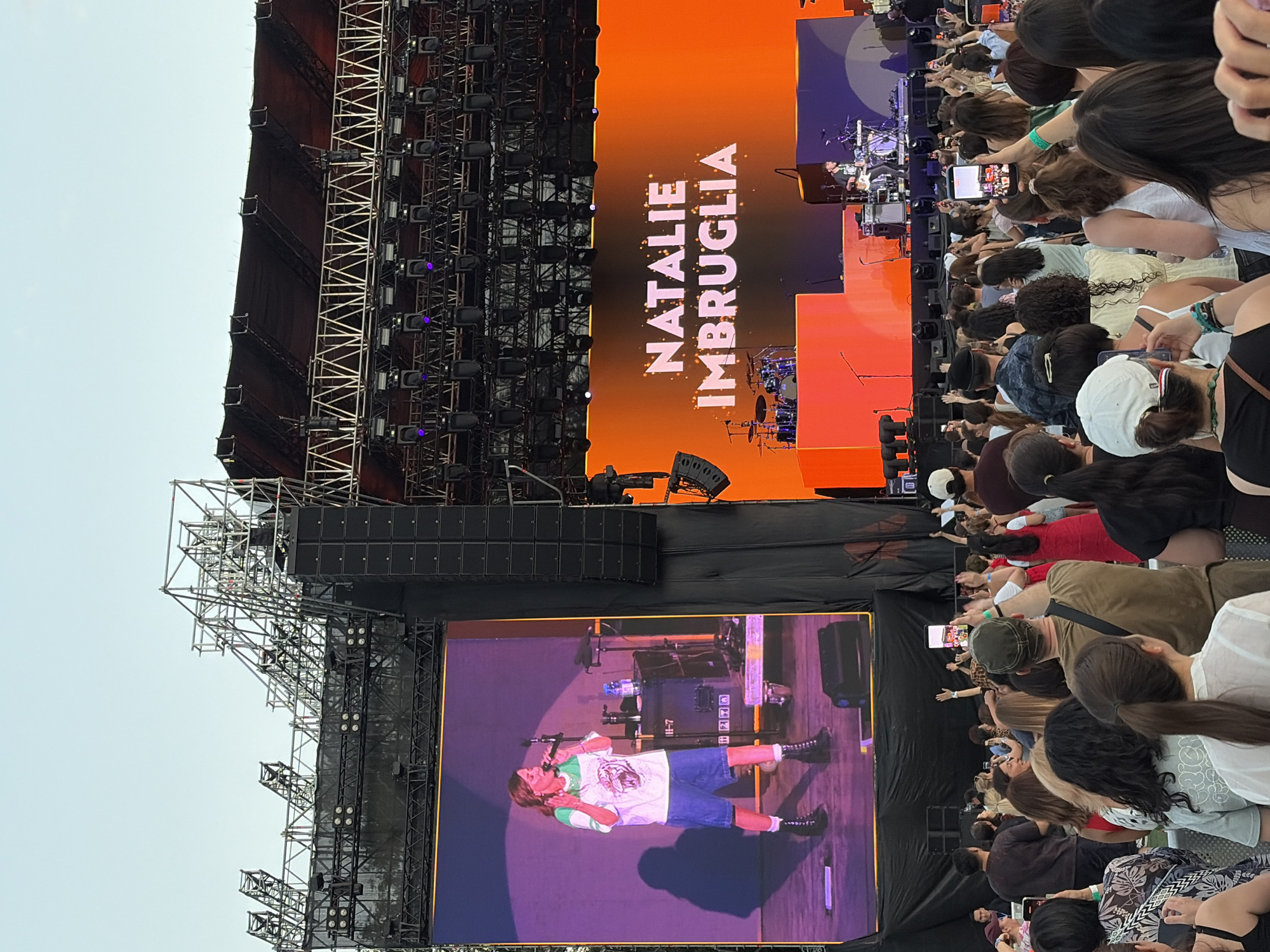The image depicts a large outdoor concert with a massive stage. A large screen on the left shows a woman singing, and a screen on the right displays the name "NATALIE IMBROGULIA" in large, bright orange letters. The stage itself has a drum set and other musical equipment visible. Below the stage, a crowd of people are gathered, many holding up their phones to record or take pictures. The overall environment appears to be a festival or concert venue with a clear sky. The image does not contain any specific visual cues that would indicate the location as Almaty, Kazakhstan.