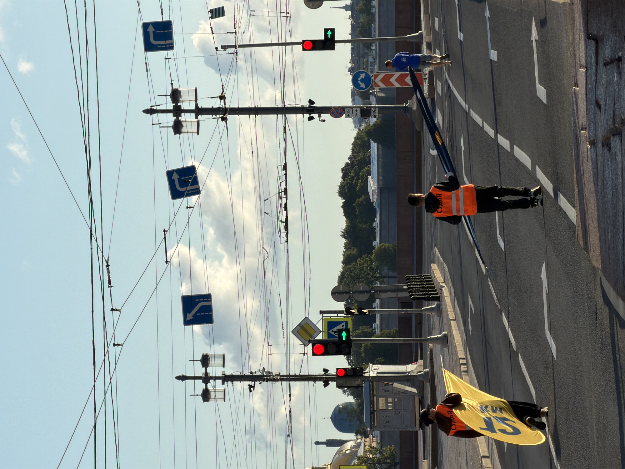 The image captures a street scene in Saint Petersburg, Russia, likely near a bridge, judging by the expanse of water visible in the background and the architectural style of the buildings. The sky is a clear blue with scattered white clouds. The foreground features a road with lane markings, a pedestrian crossing, and a sidewalk.
Two individuals are central to the scene, both wearing high-visibility orange vests. The person on the left is walking towards the viewer's left, holding a large yellow sign with blue lettering. The lettering is partially obscured by the sign's curvature and lighting, but appears to be Cyrillic. This individual is also wearing a dark baseball cap and dark pants.
The second person, to the right, is walking away from the viewer, holding a long, dark, rectangular object horizontally, possibly a barrier or directional sign. This person is wearing a blue jacket and grey shorts.
Several traffic lights are visible, with one on the left displaying red lights, and another further down the road showing a red light and a green arrow. A pedestrian crossing signal shows a green walking figure. Overhead, a complex network of electrical wires, likely for trams or trolleybuses, crisscrosses the sky, supported by lampposts.
In the middle ground, behind the road, are a row of lampposts and some temporary structures that might be related to roadwork or events. Beyond that, trees and buildings line the horizon. The iconic blue dome of a mosque or religious building is visible in the far left background. The overall atmosphere suggests an organized event or traffic management in progress.