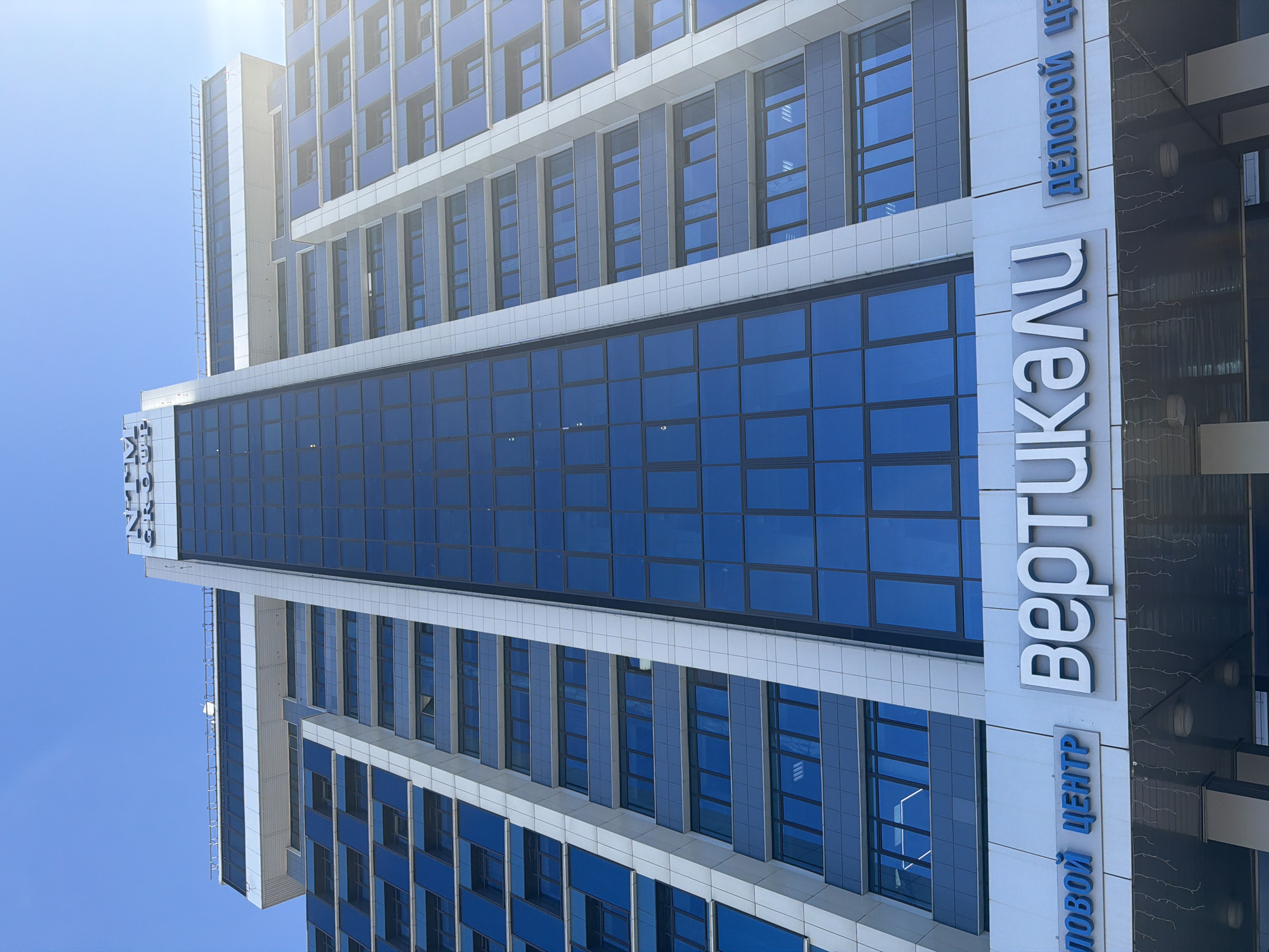 The image shows the exterior of a modern, high-rise office building, captured from a low angle looking upward. The building features a blue and white facade with numerous rectangular windows. A prominent vertical sign displays the word "ВЕРТИКАЛИ" (VERTICALLY) in large white letters, and below it, "ДЕЛОВОЙ ЦЕНТР" (BUSINESS CENTER) in blue Cyrillic. Based on the Cyrillic text, the building is located in a Russian-speaking area. Given the presence of the sign "VERTICALI" and "BUSINESS CENTER" it is probably in Krasnoyarsk, Russia. The sky is clear and blue. No people are visible. The photograph focuses on the architecture and branding of the building.