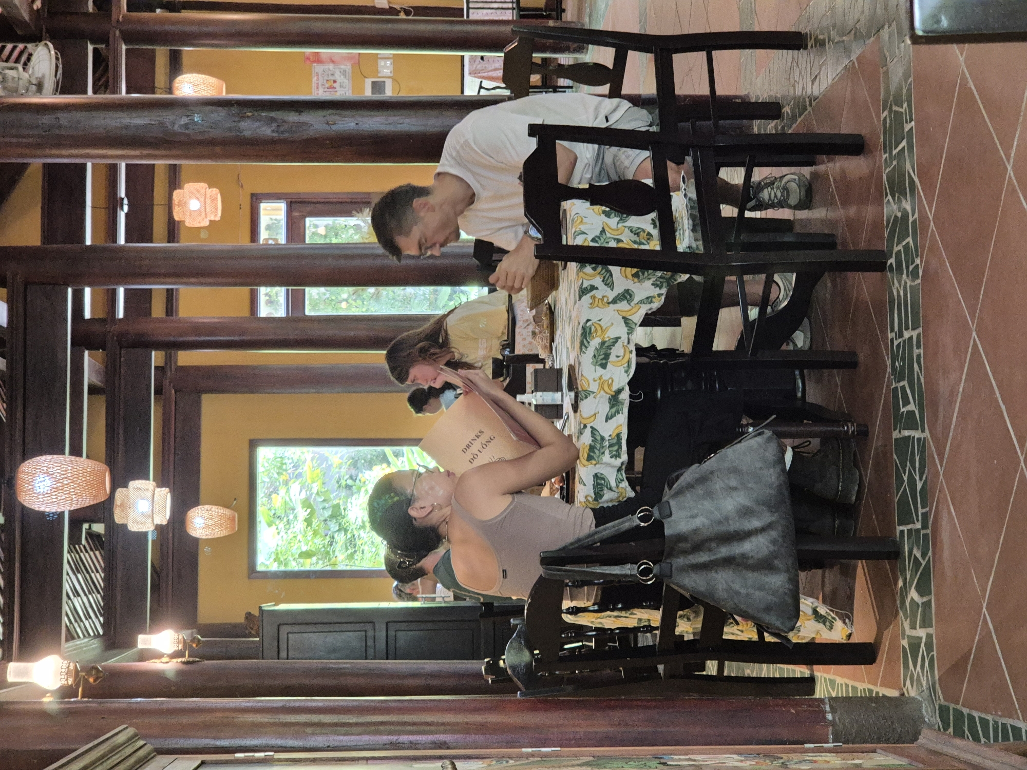 The image depicts an indoor dining area at the Han River Restaurant in Da Nang, Vietnam. The restaurant features traditional wooden architecture with exposed beams and pillars. Several people are seated at tables, engaged in eating and conversing. One woman, wearing glasses and a brown tank top, is reviewing a menu. A man in a white t-shirt and shorts sits opposite her. Another woman with long hair is visible in the background, also seated at a table. The tables are covered with a tablecloth adorned with a banana leaf pattern. Various lamps, including woven pendant lights and wall sconces, illuminate the space. The floor is tiled with reddish-brown and green tiles. The overall ambiance suggests a casual dining experience in a culturally rich setting.