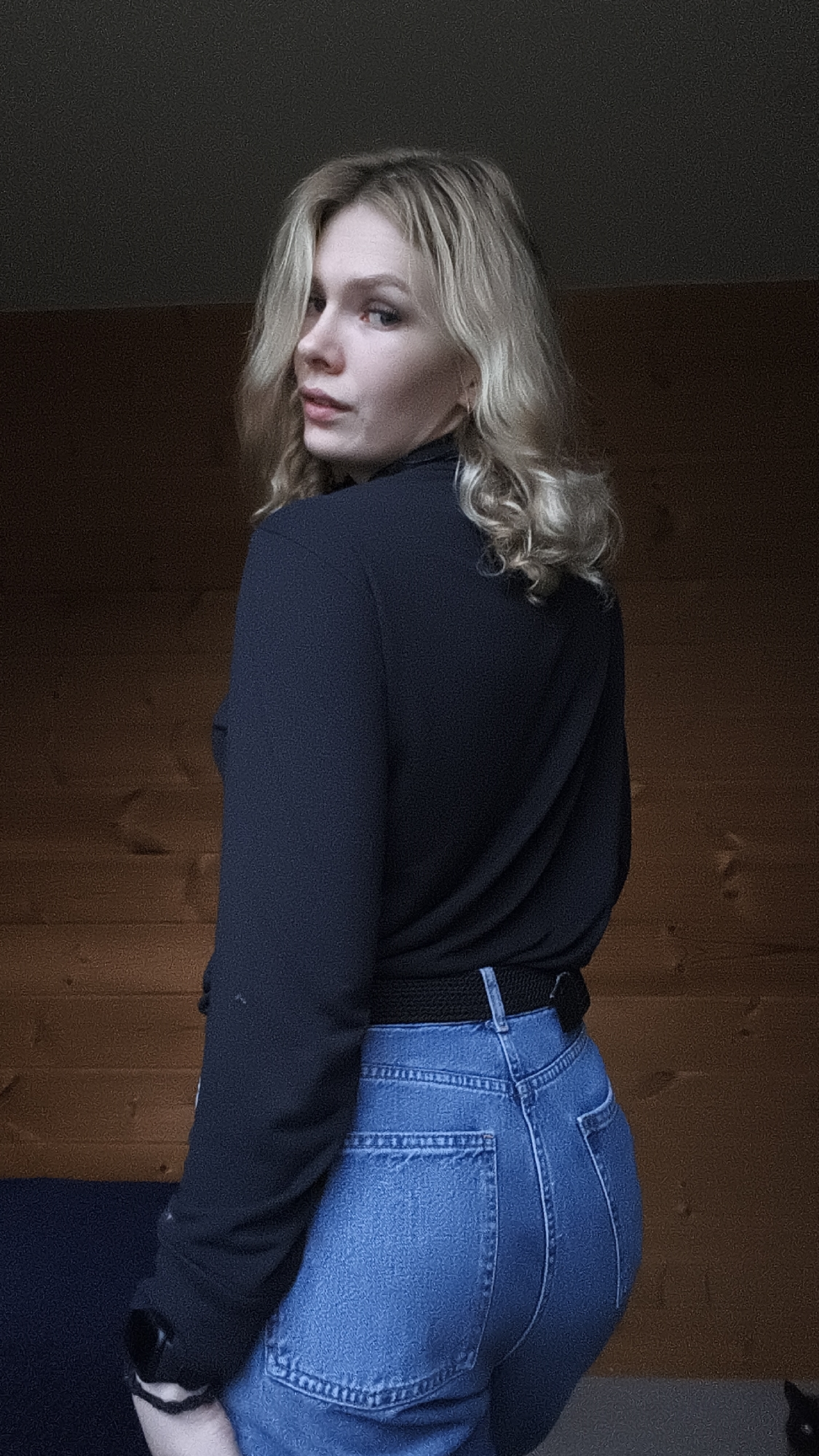 A young woman with blonde, shoulder-length hair is posing indoors. She is turned slightly to her right, looking back over her shoulder towards the viewer. She is wearing a long-sleeved black top and blue jeans with a black belt. The background is a brown wall, possibly wood paneling, with a darker area above. There's also a glimpse of a dark blue surface in the lower left corner. The specific location is Русский Вожой, Russia.